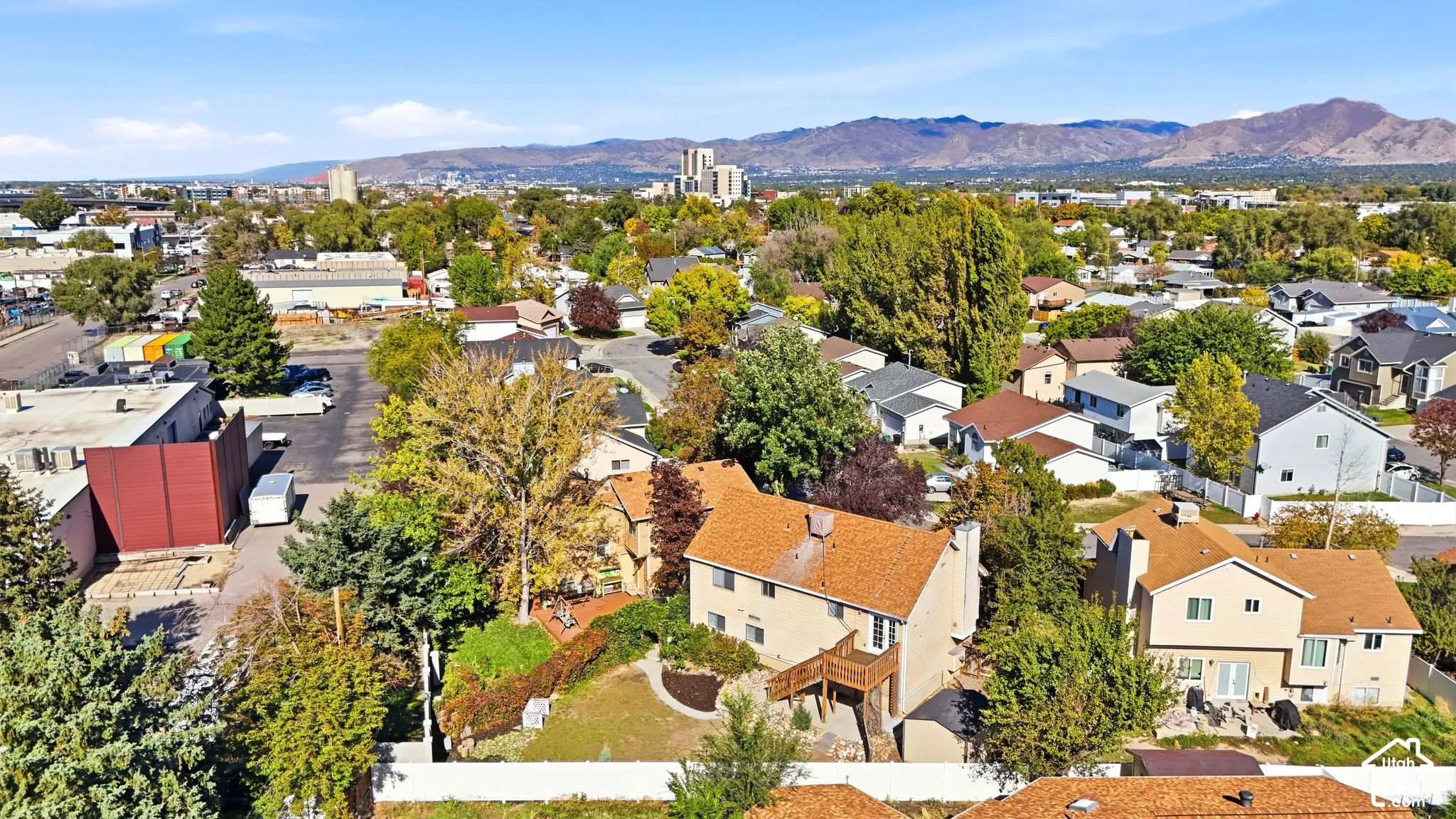 Aerial perspective featuring a multi-level residence with a wood-composite deck, mountain backdrop, mature trees, and fenced grounds