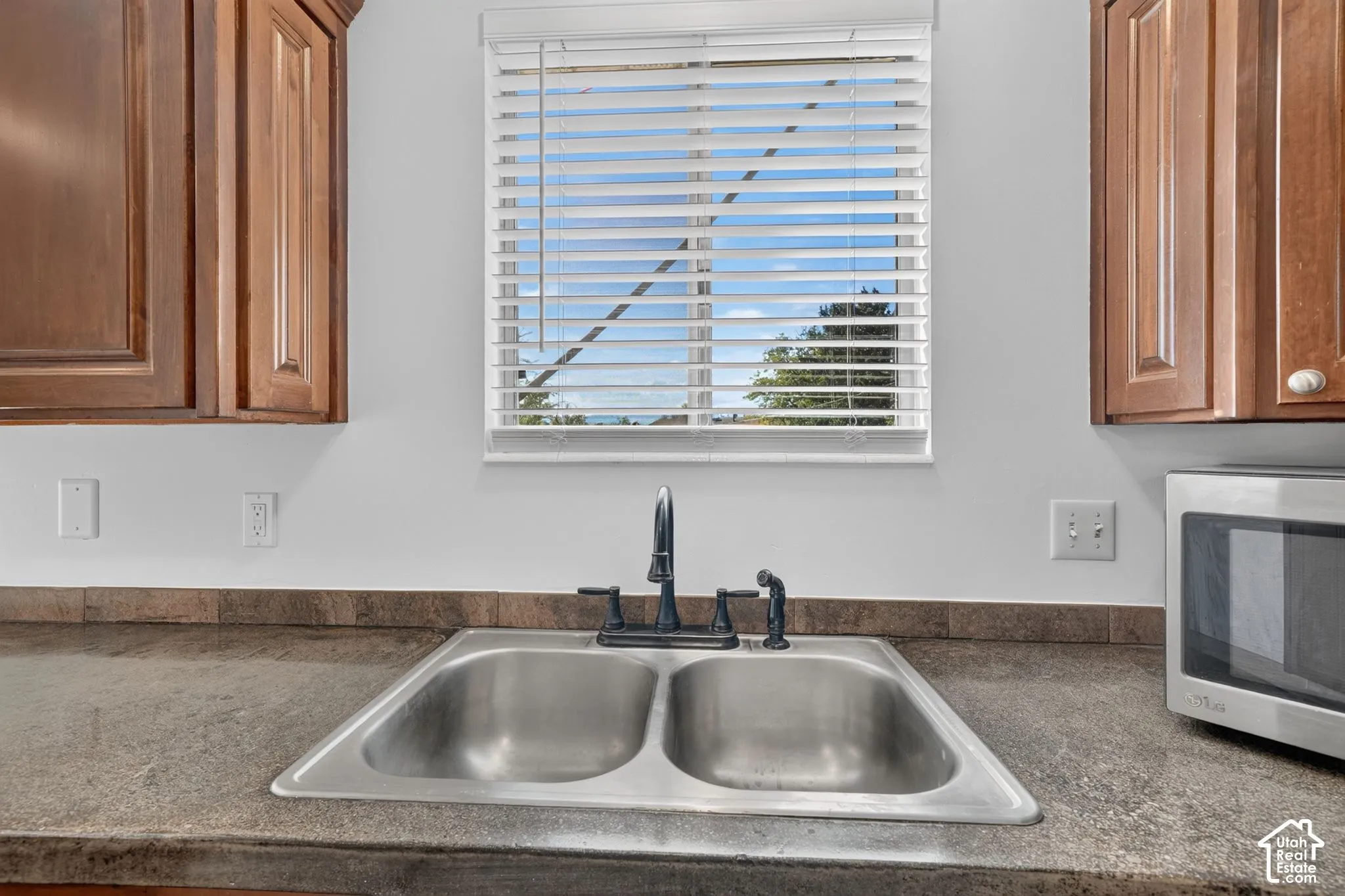 Stainless steel dual-basin undermount sink with a dark-finish faucet, stone-surfaced countertop, tiled backsplash, and wood-finish cabinetry