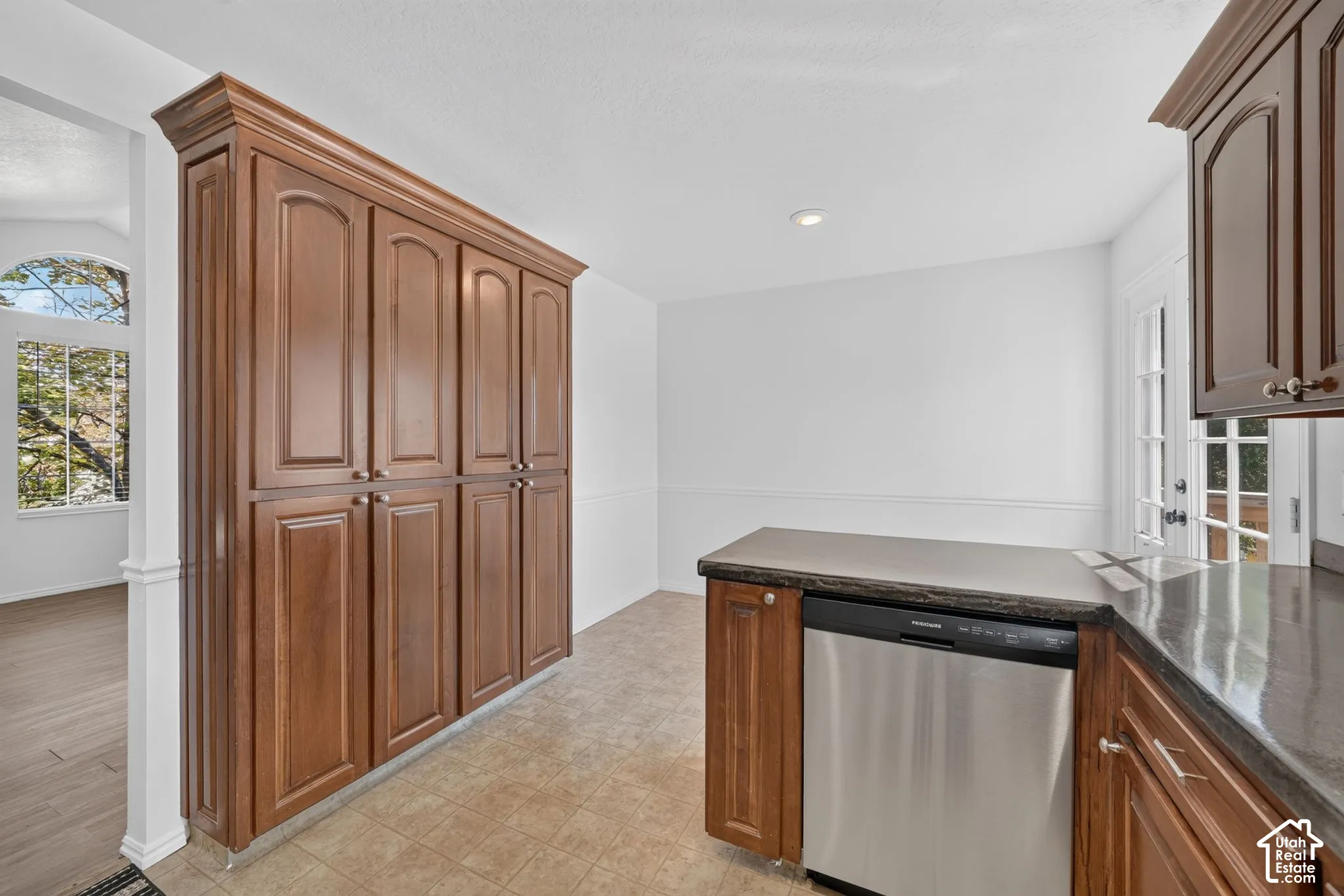 Floor-to-ceiling wooden cabinetry, stainless steel dishwasher, dark stone-finish countertops, tile flooring, and arched window