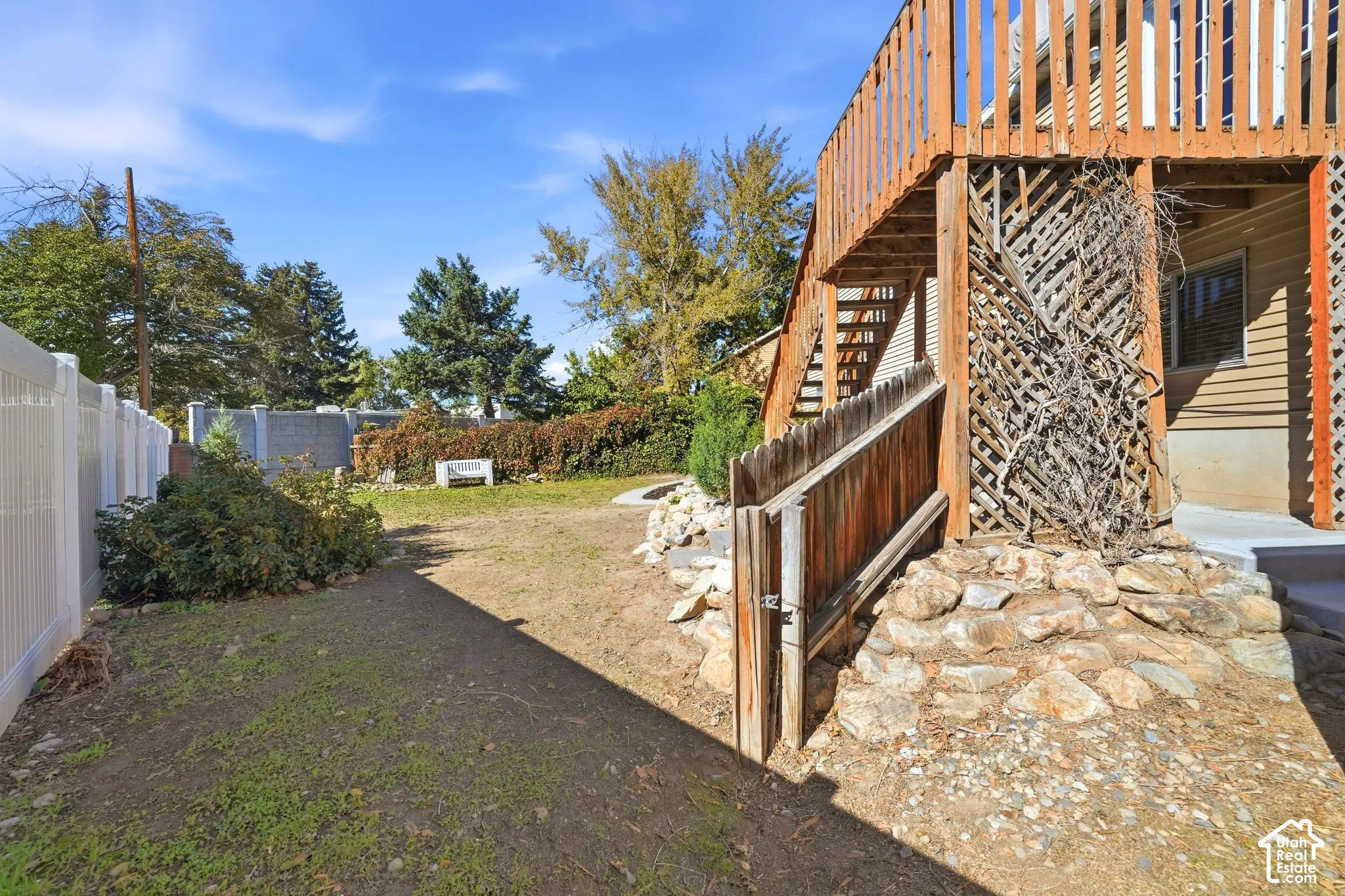 Backyard featuring elevated wood deck, stone hardscaping, lattice privacy screening, and white vinyl perimeter fencing