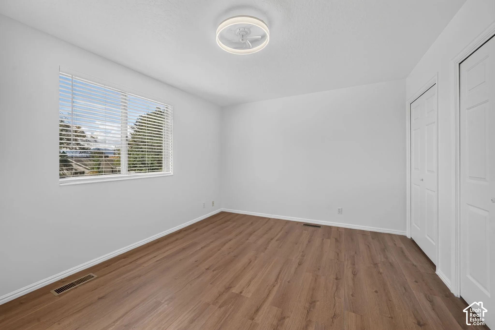 Neutral-toned room featuring wood-finish flooring, a large window with horizontal blinds, a circular flush-mount ceiling light fixture, and double-door closet cabinetry
