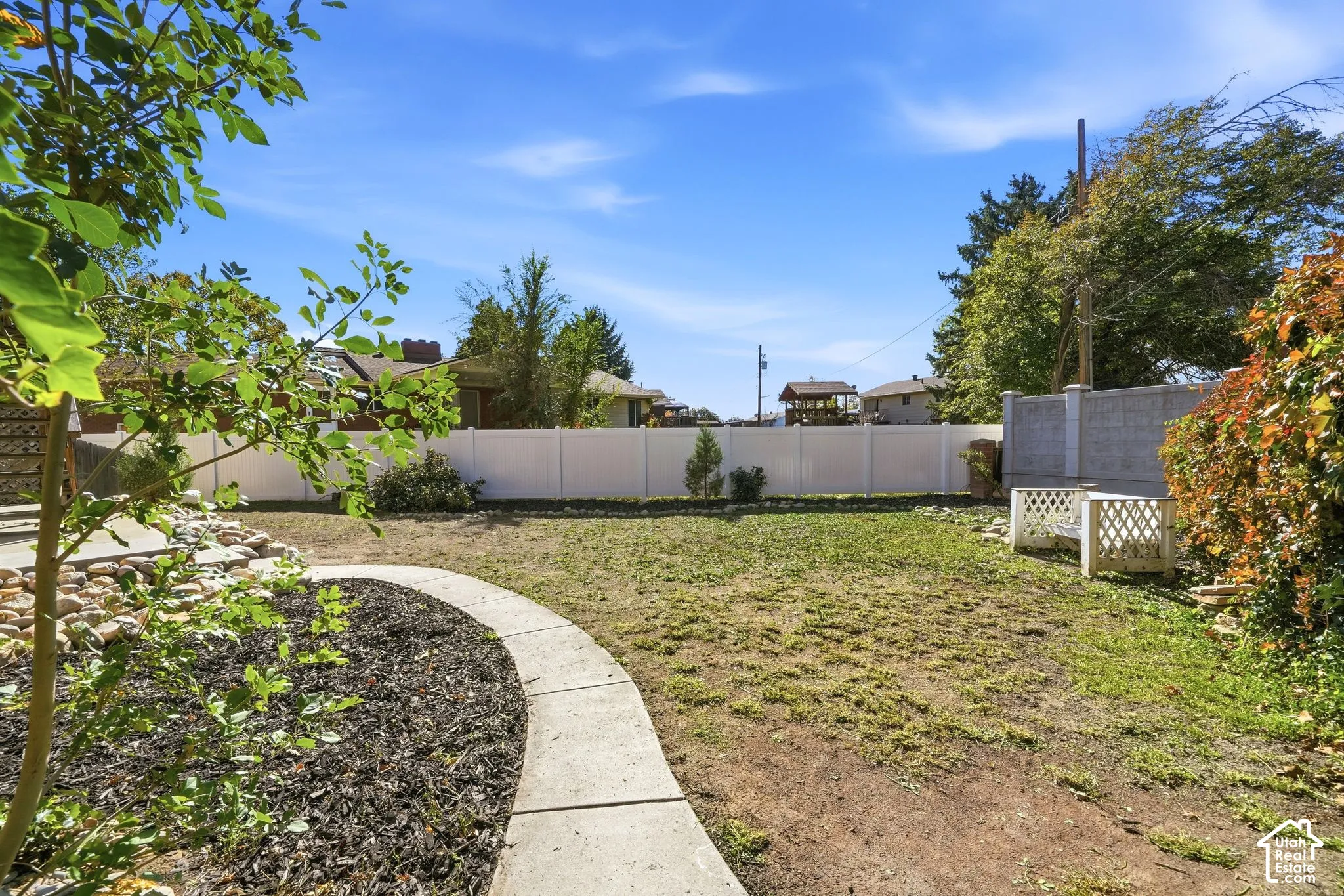 Backyard featuring a curved concrete walkway, stone landscaping border, vinyl perimeter fencing, and mature trees