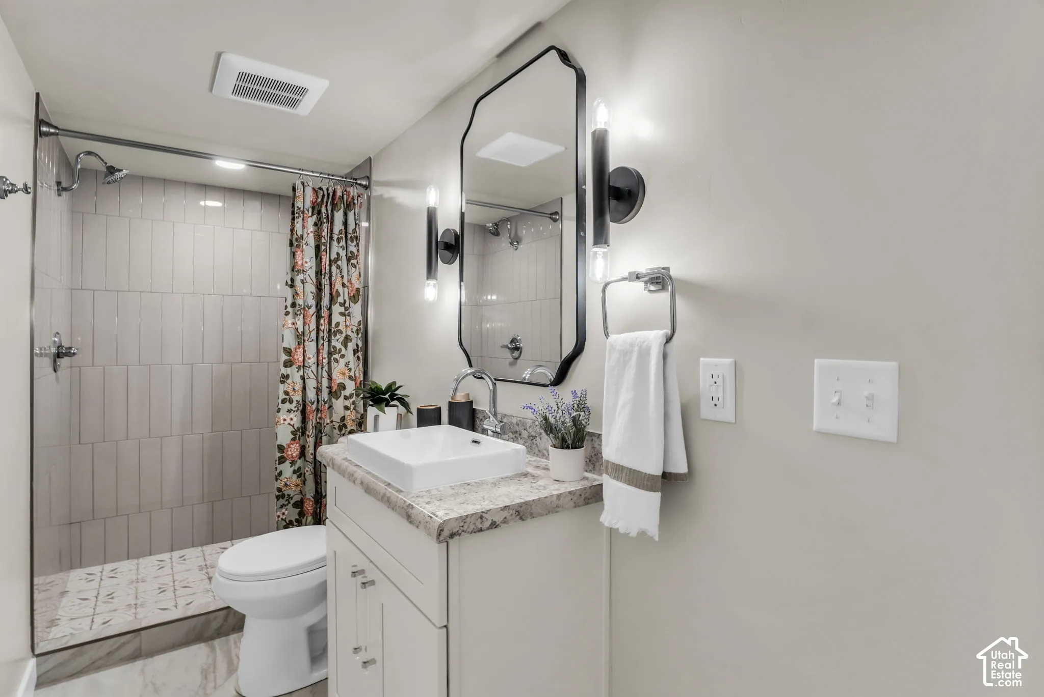 Bathroom featuring a vanity with a surface-mounted sink, a large wall-mounted mirror, modern wall sconces, a shower with vertical tile, and a patterned shower floor