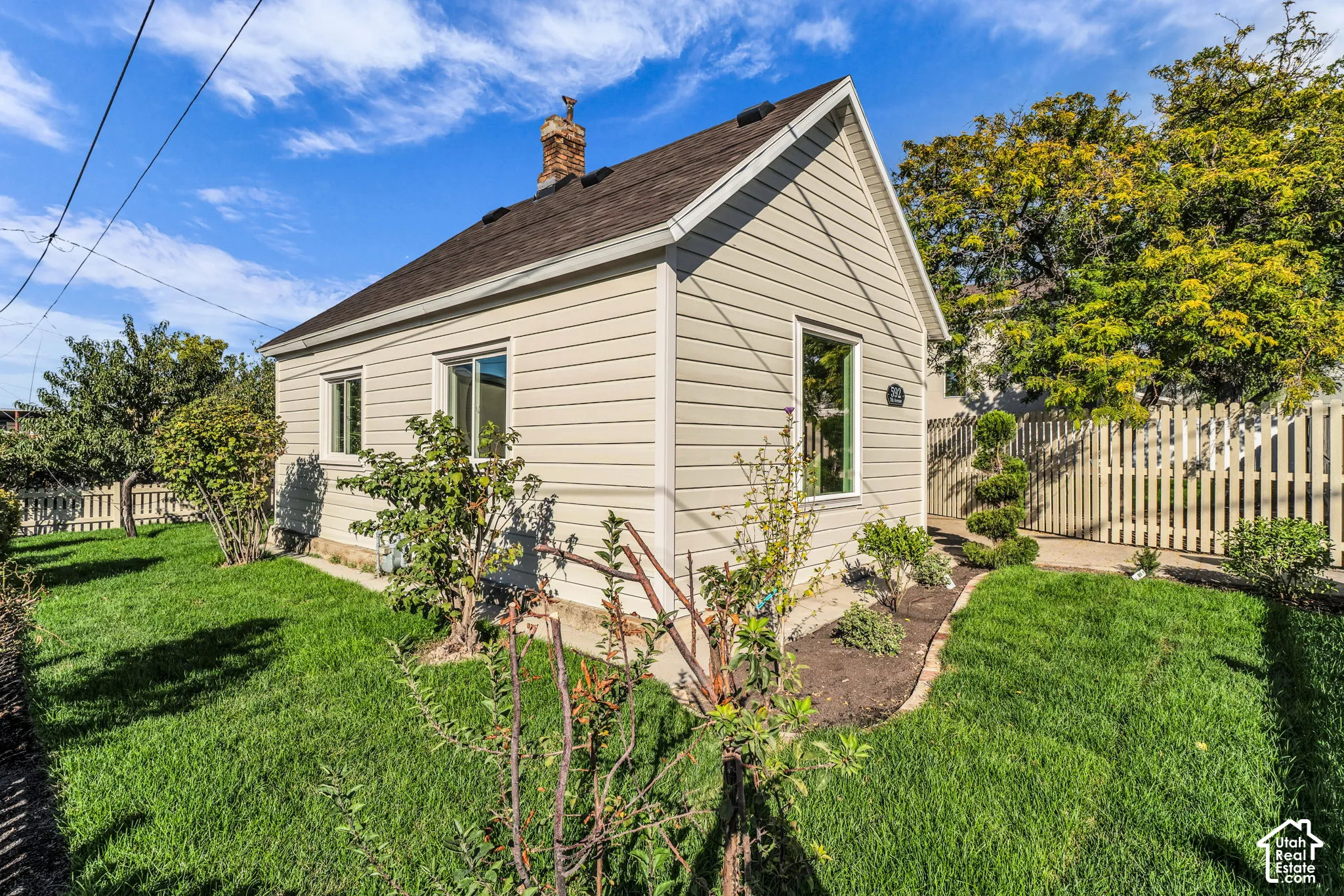 Gable roofline, light-colored siding, multiple windows, brick chimney, and a white picket fence