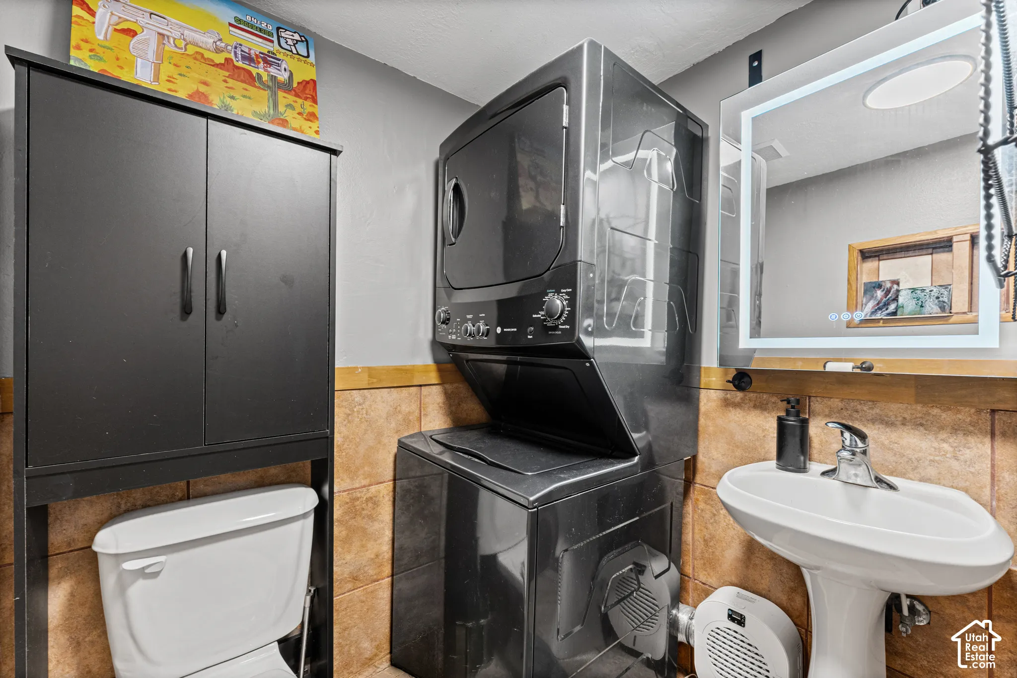 Stacked washer and dryer unit, porcelain pedestal sink with polished chrome faucet, and integrated LED-backlit mirror, finished with tile wall accents and built-in vertical storage cabinetry