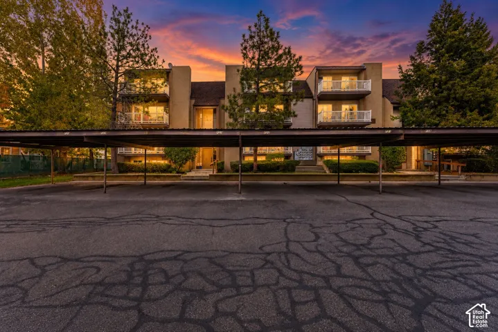 Multi-level building featuring stucco siding, metal-frame carport parking, private balconies with white railings, and mature evergreen landscaping