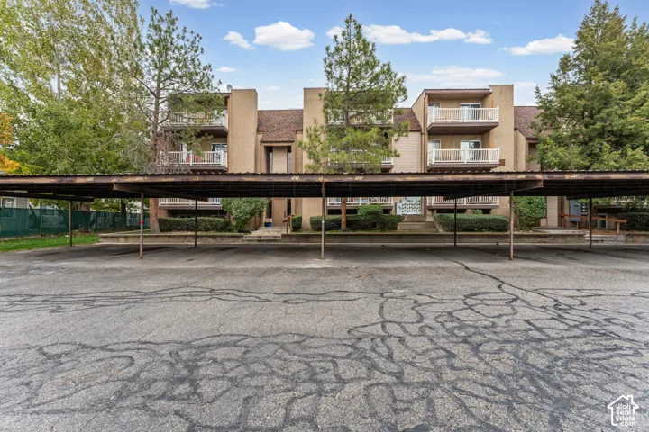 Multi-story building featuring stucco siding, individual private balconies with white railings, and an expansive paved surface lot with covered parking structures
