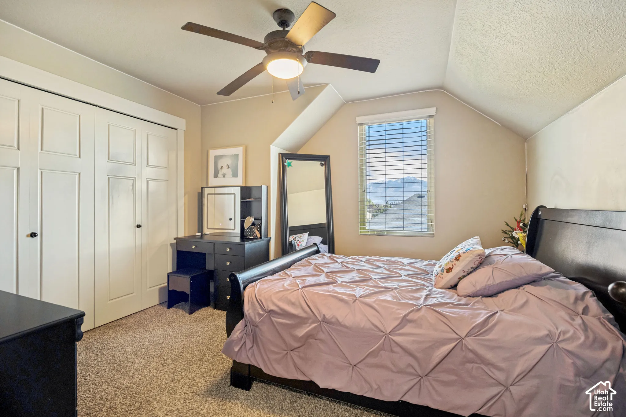 Neutral-toned room featuring a vaulted ceiling, dual-door closet with paneled detailing, central ceiling fan with integrated light fixture, carpeted flooring, and a window with mountain backdrop