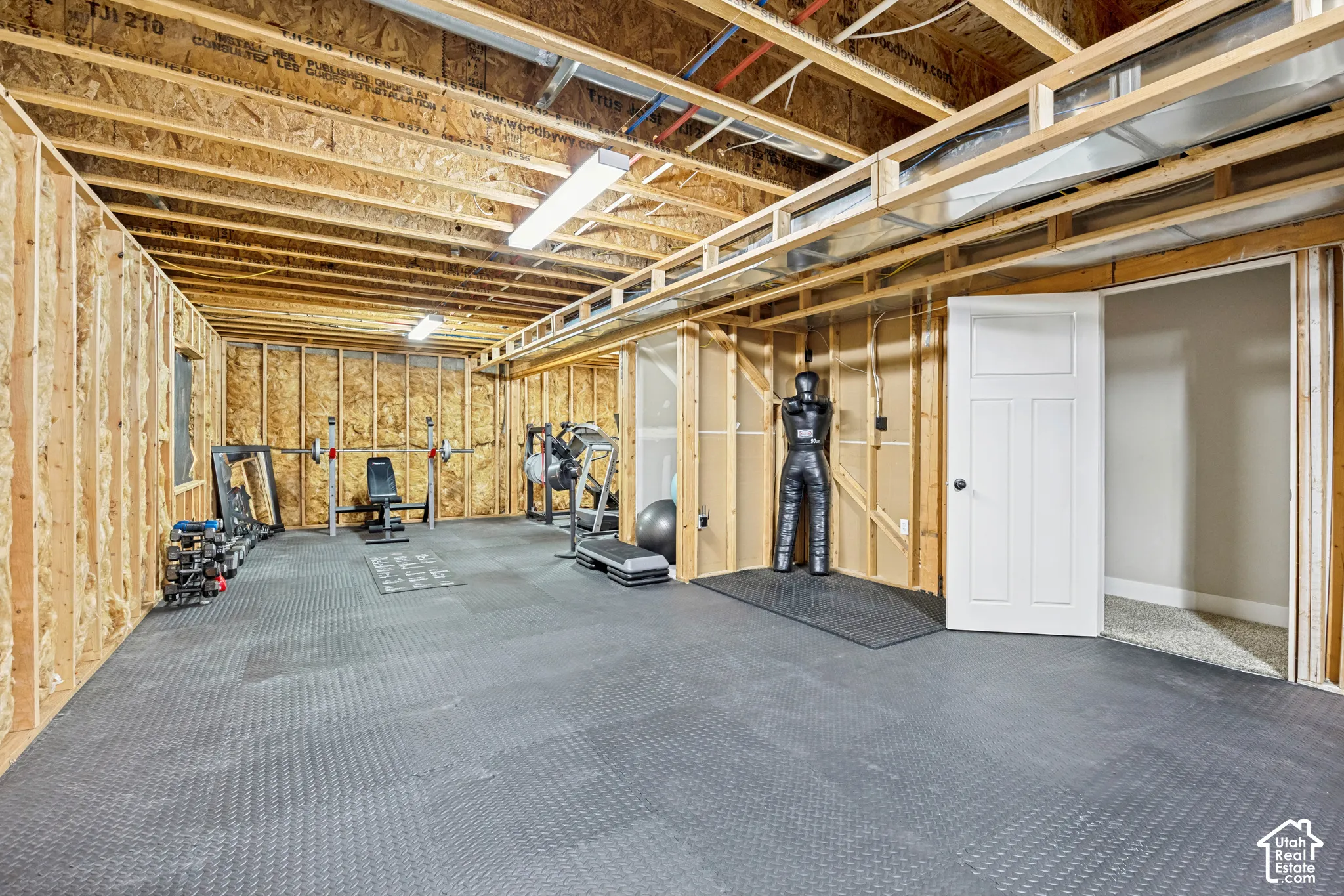 Unfinished utility space featuring exposed wood-stud wall framing, open joist ceiling, rubberized interlocking flooring, and a white interior door