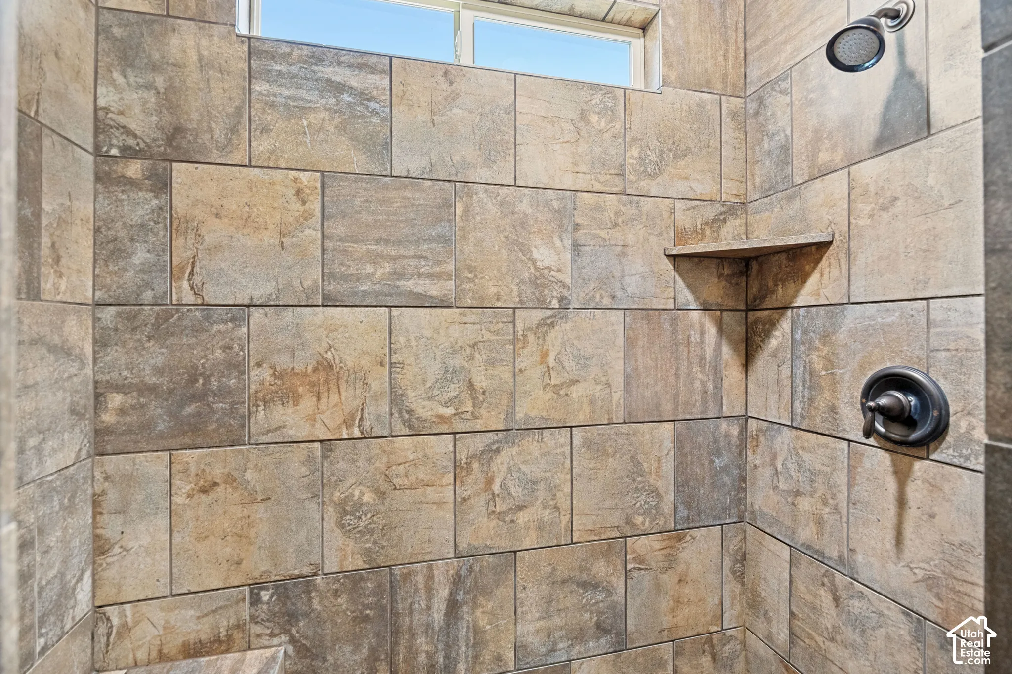 Shower featuring large-format ceramic wall tile, a built-in corner shelf, horizontal transom window, and oil-rubbed bronze plumbing fixtures