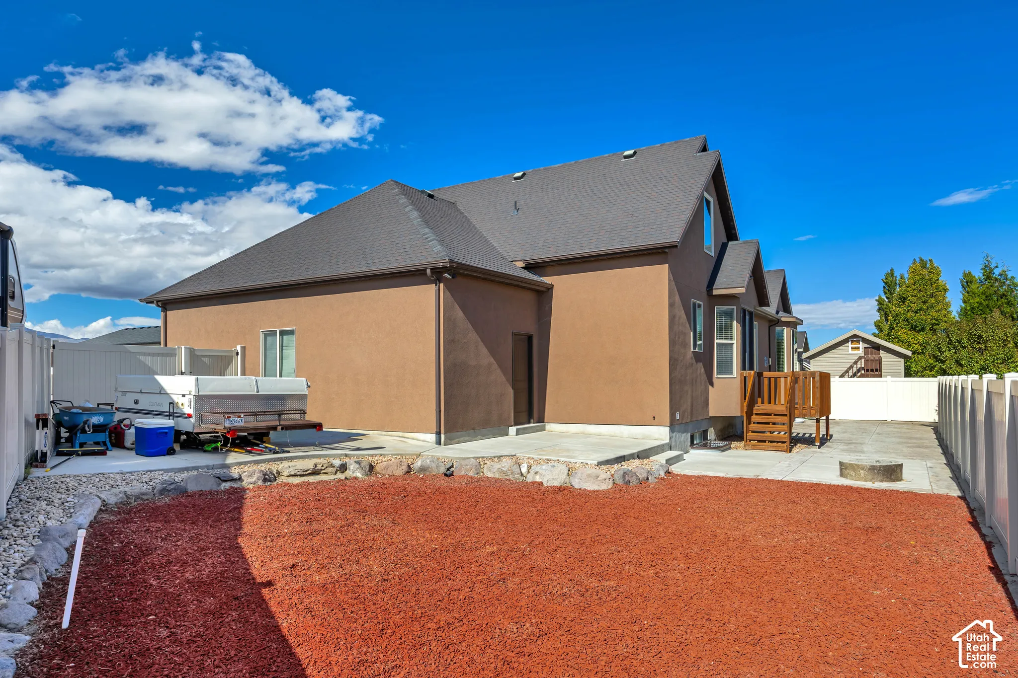 Exterior featuring a brown stucco finish, asphalt shingle roof, concrete patio, wood deck, and perimeter vinyl fencing