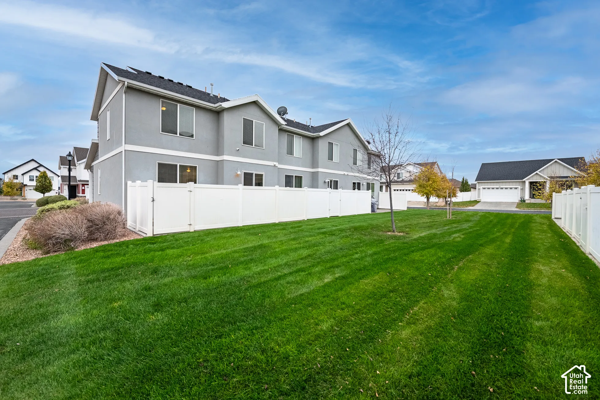 Multi-unit building exterior featuring grey siding, a gabled roofline, dual-tone window frames, a vinyl privacy fence, and an expansive lawn