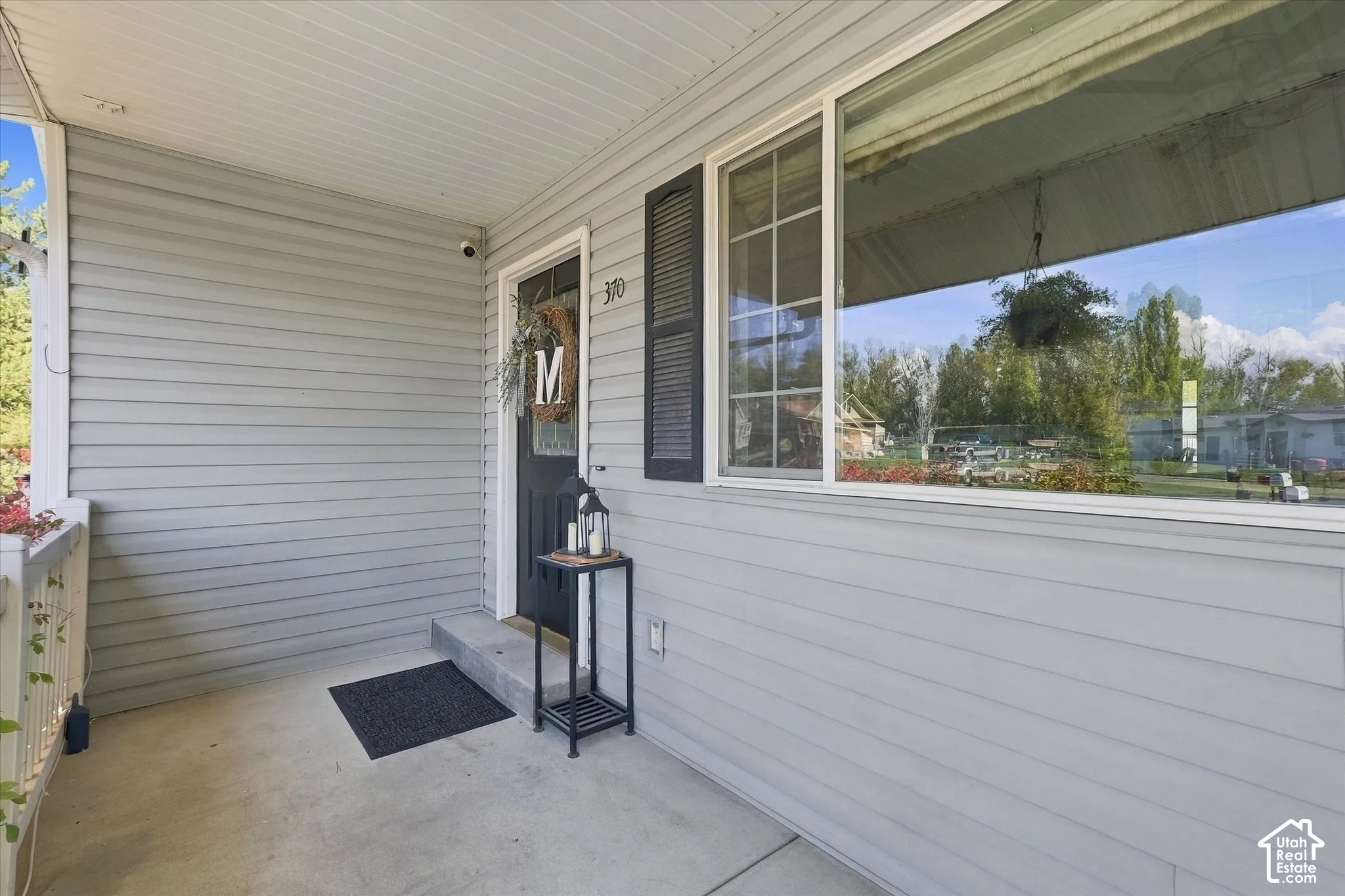 Covered front porch featuring light grey horizontal siding, a paneled exterior door with glass inlay, a large multi-pane window with dark shutters, and a concrete floor