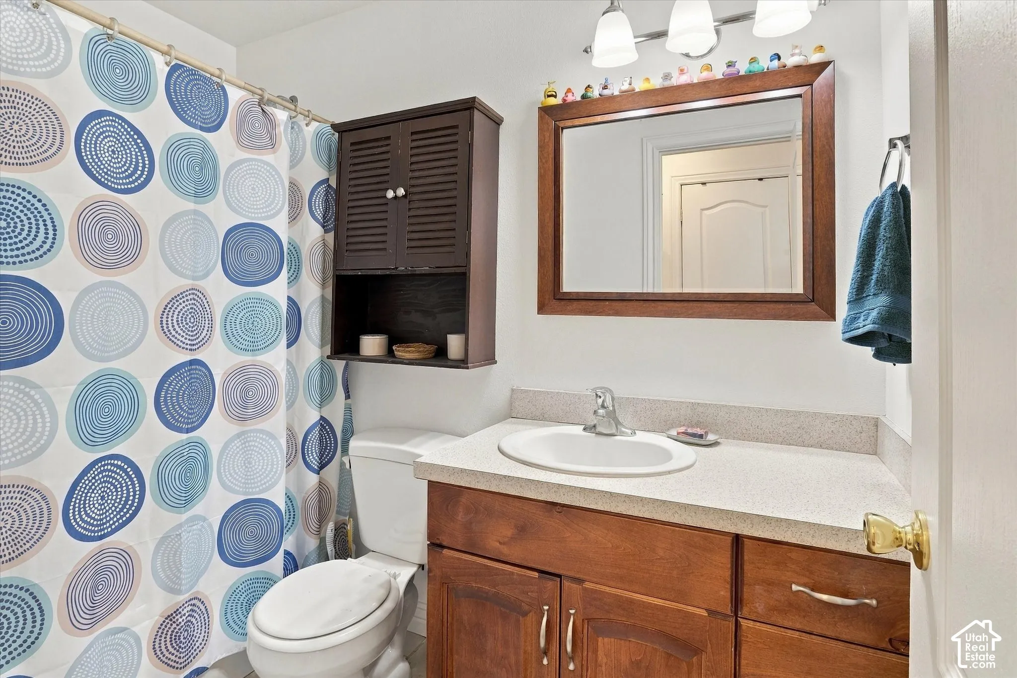 Bathroom featuring a wood-finish vanity with a composite countertop and integrated basin, wall-mounted storage cabinetry, a framed vanity mirror, and three-light overhead vanity fixture