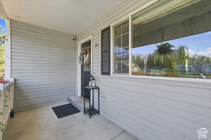 Covered front porch featuring light grey horizontal siding, a paneled exterior door with glass inlay, a large multi-pane window with dark shutters, and a concrete floor