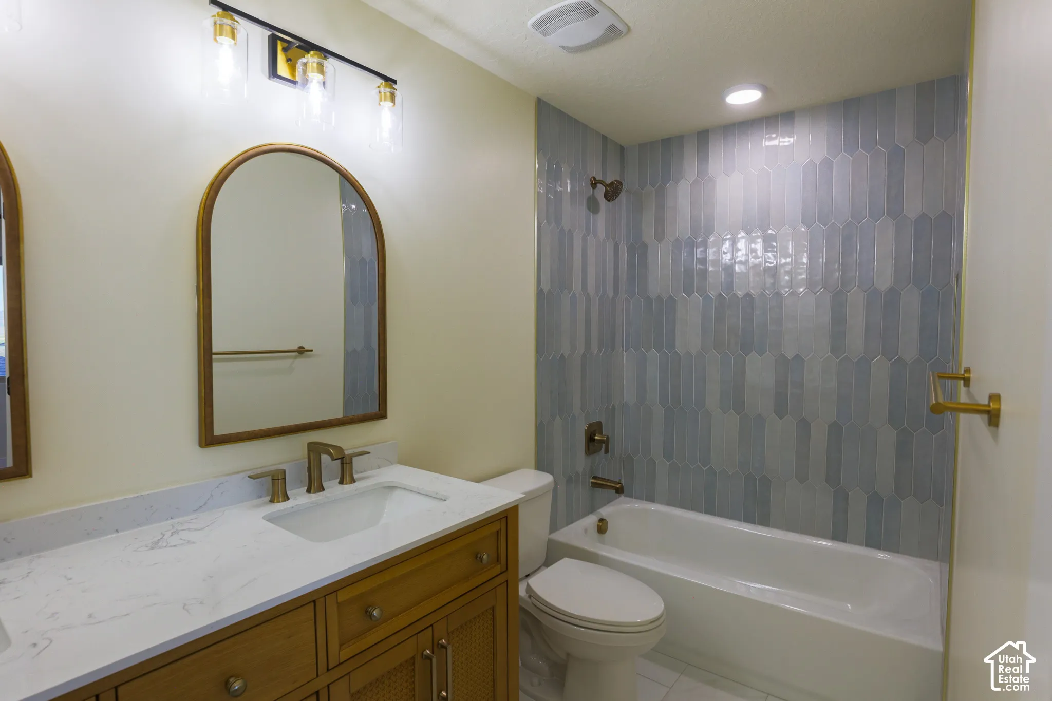 Bathroom featuring a vanity with a stone countertop, an arched mirror, three-light vanity fixture, and a tub surround with patterned vertical tile