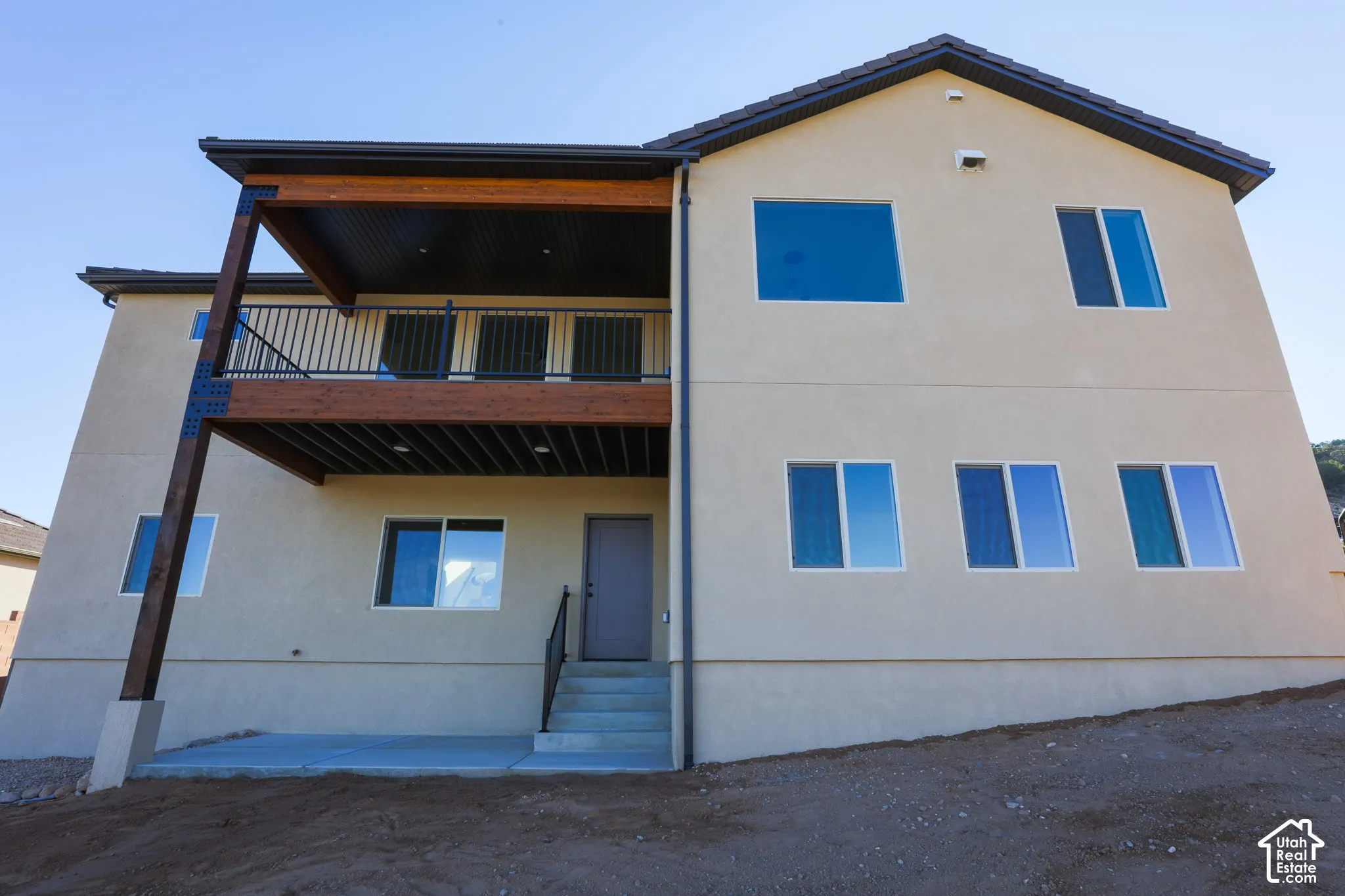 Stucco exterior featuring an elevated wood-beam balcony, metal railing, concrete patio, and multiple windows with white trim
