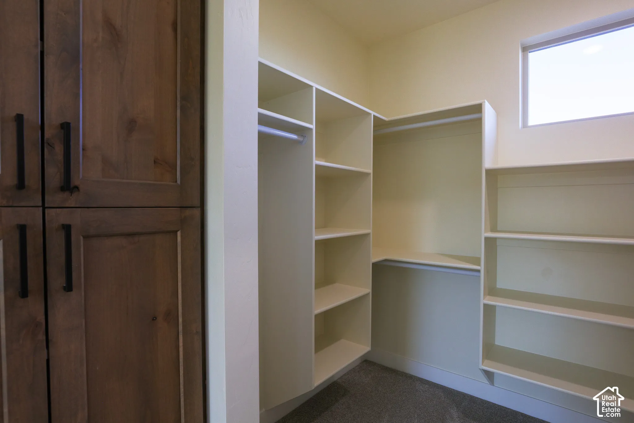 Walk-in storage space featuring built-in white shelving, integrated hanging rods, floor-to-ceiling cabinetry with dark hardware, and a high-set window