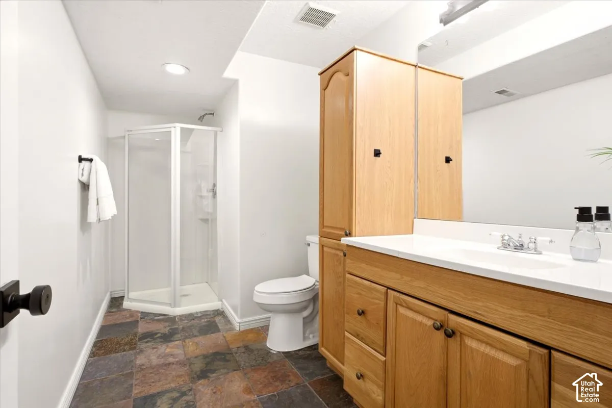 Bathroom featuring natural stone tile flooring, corner glass-enclosed shower, light-toned wood cabinetry with built-in storage tower, white solid-surface countertop, and wall-to-wall mirror