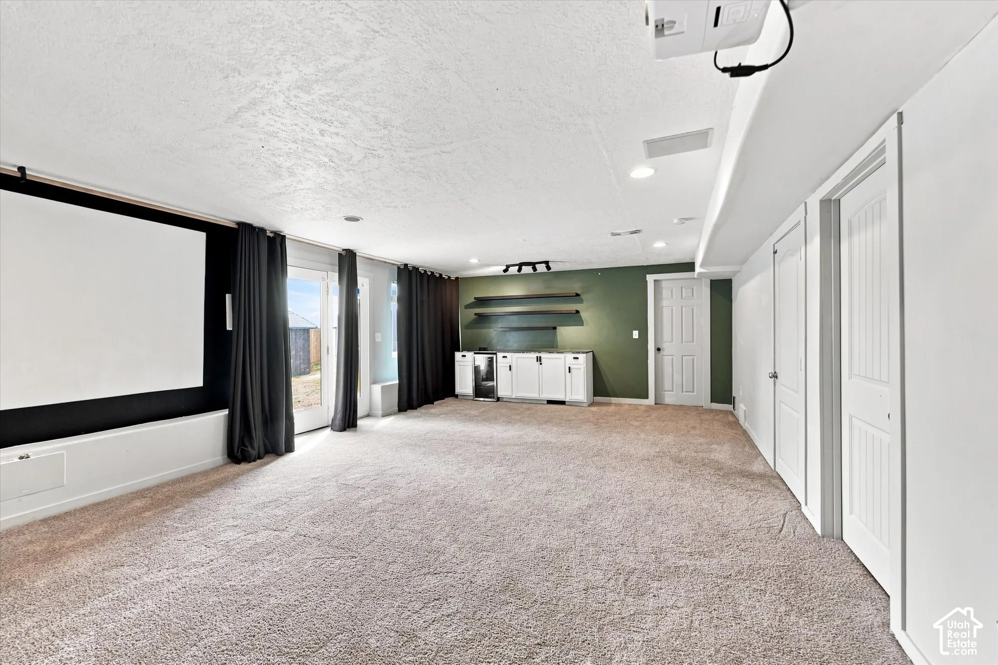 Built-in projection screen, wet bar featuring white cabinetry and stainless-steel under-counter refrigerator, wall-mounted floating shelving, recessed ceiling lighting, and glass-paneled exterior door