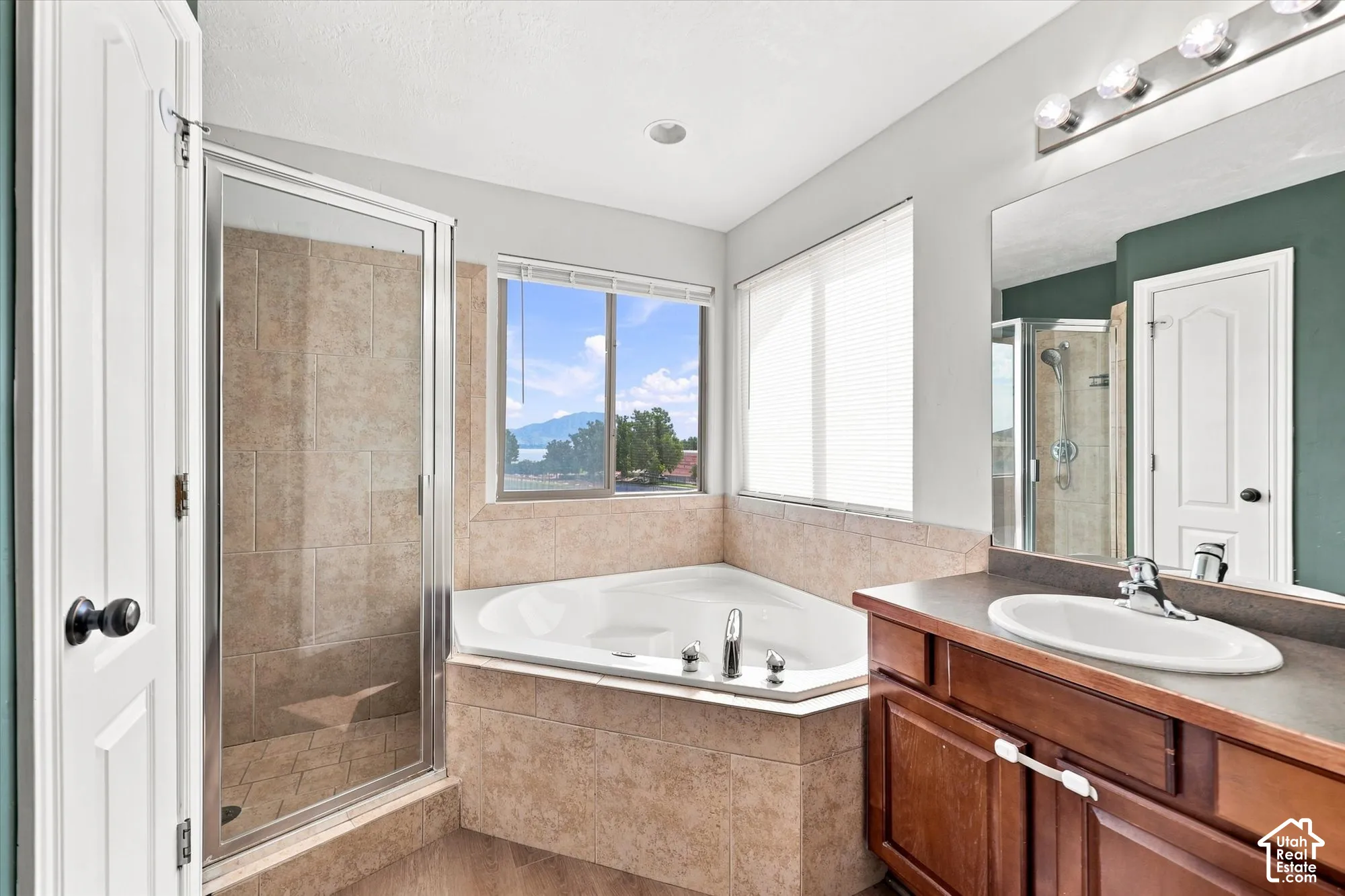 Corner soaking tub with tile surround, glass-enclosed shower, wood-finish cabinetry, surface-mounted vanity mirror, and windows featuring mountain scenery