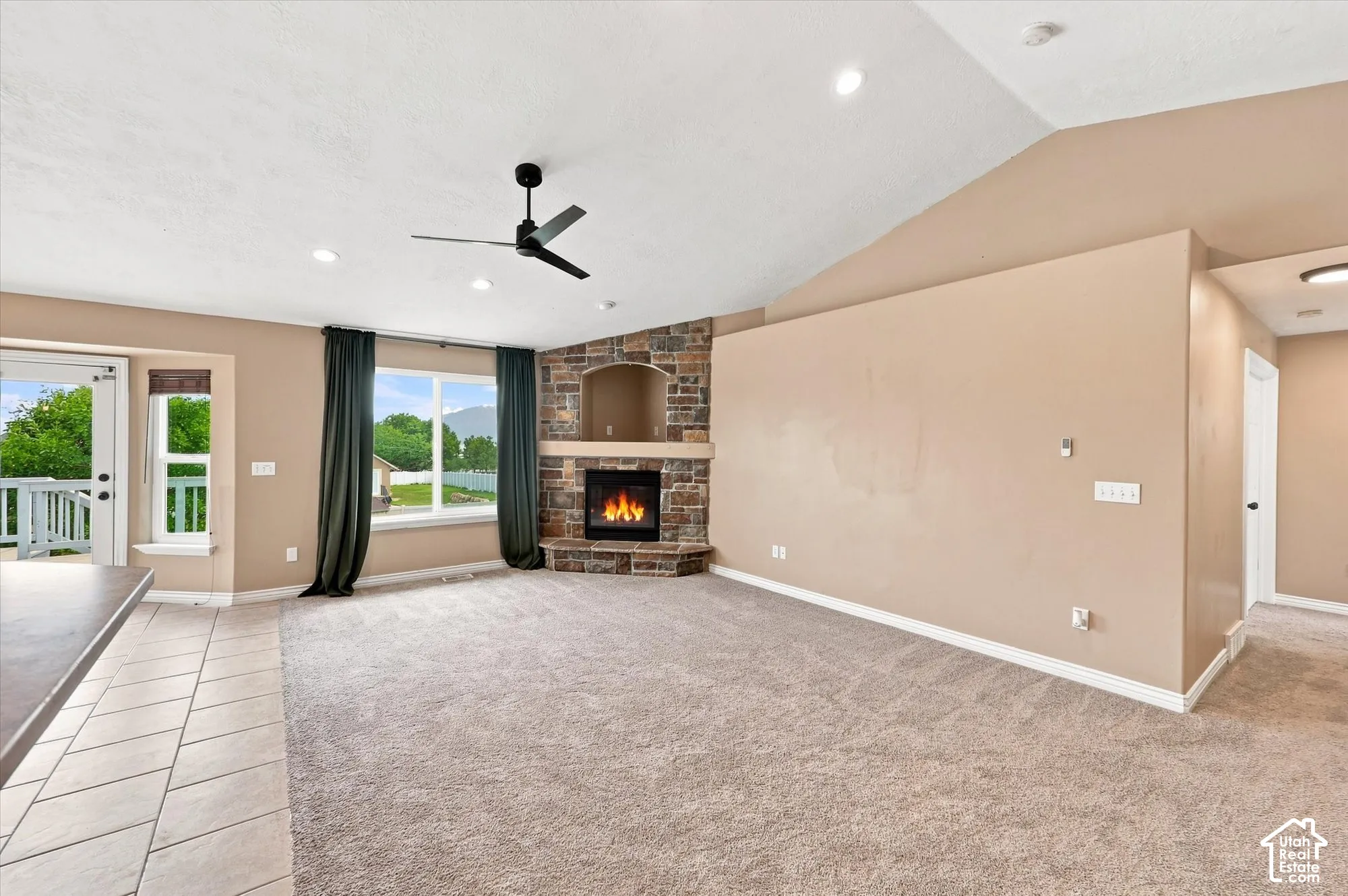 Vaulted ceiling featuring a corner stone-surround fireplace with a recessed media niche, neutral carpeting, tile flooring, and a ceiling fan