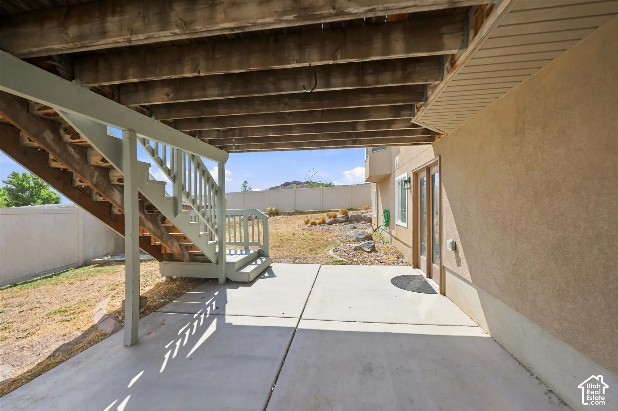 Covered concrete patio featuring a wooden staircase, stucco exterior finish, and fenced backyard with stone landscaping accents