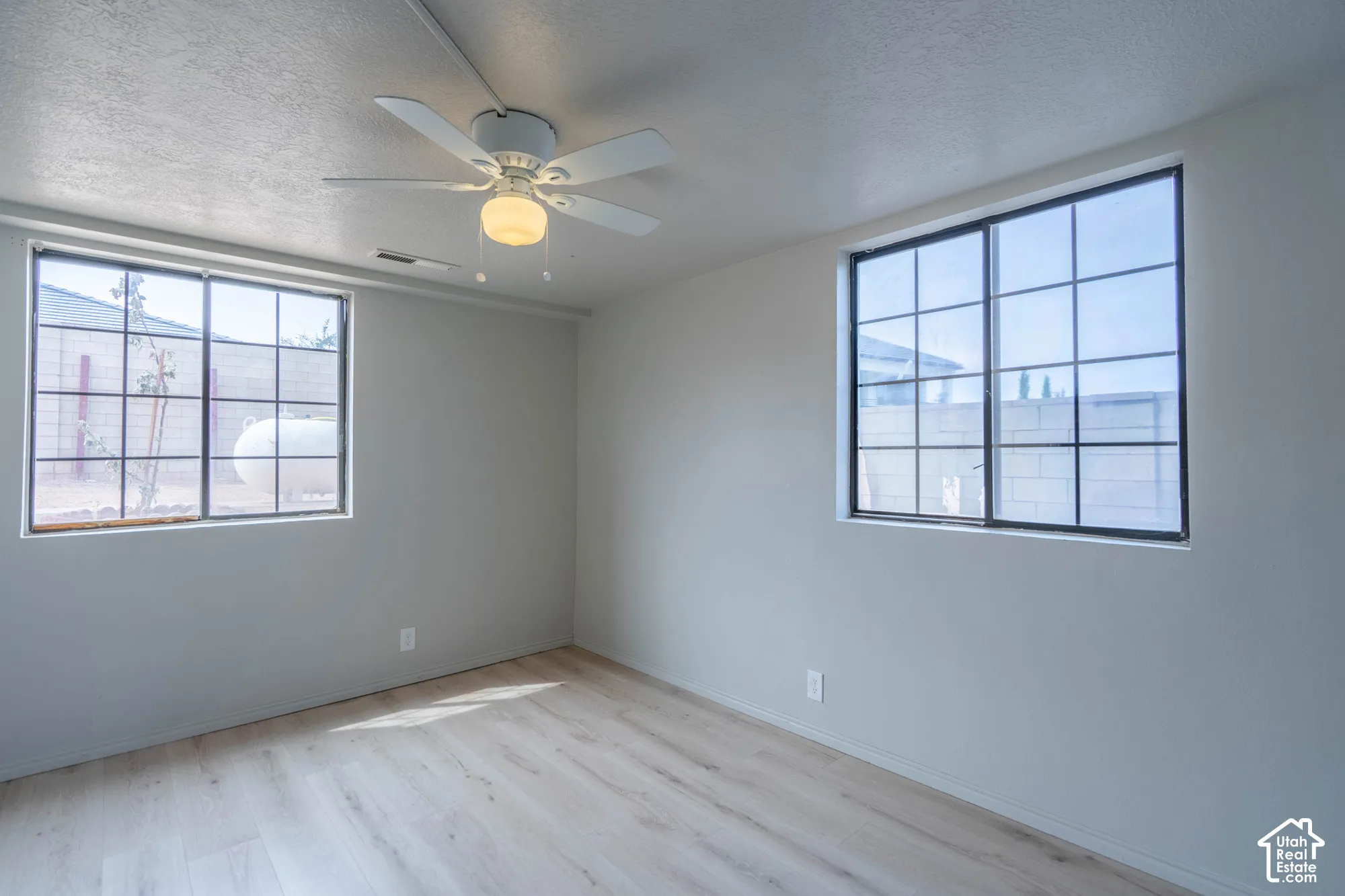 Light wood-finish flooring, two large grid-pattern windows, and a ceiling fan with integrated lighting