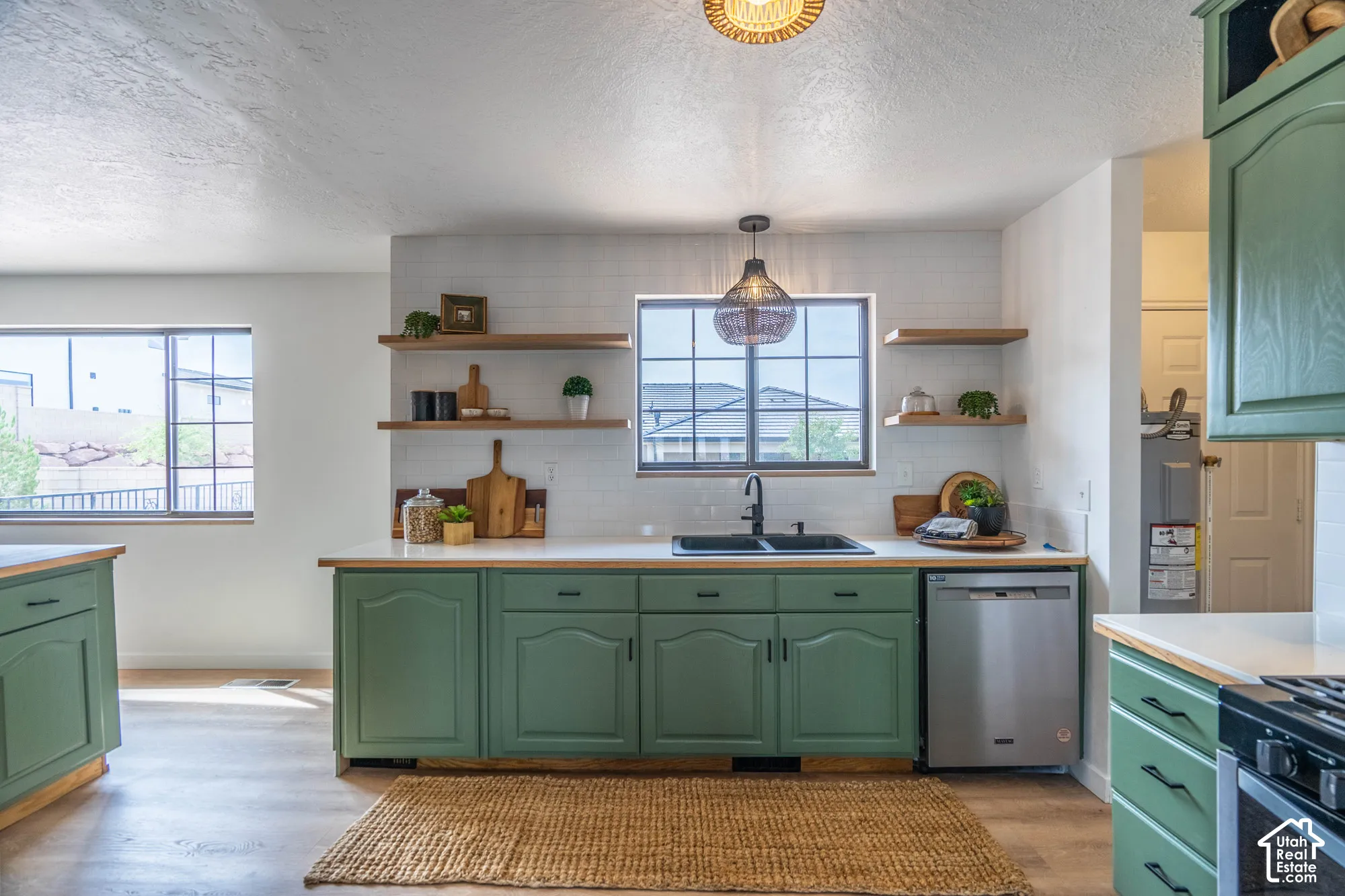 Kitchen featuring green cabinetry, light-toned countertops, a stainless steel dishwasher, a black sink with a matching faucet, and open wood-finish shelving