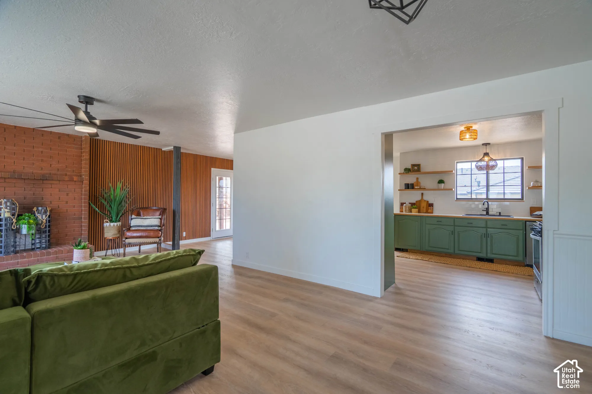 Light wood-finish flooring throughout, brick fireplace surround, wood slat accent wall, kitchen with green cabinetry and open shelving, modern ceiling fan