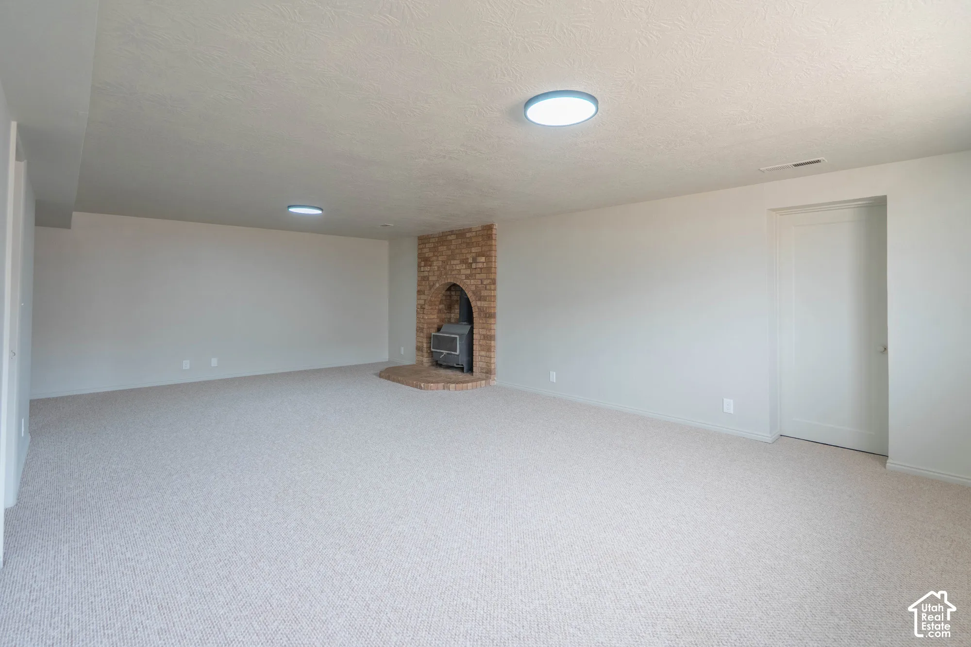Expansive interior space with light-colored carpeting, a brick-clad fireplace with a wood-burning insert, and multiple flush-mount ceiling lights
