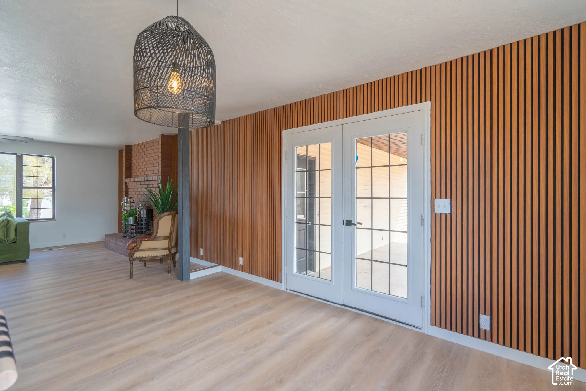 Light wood-finish flooring, accent wall with vertical wood slat panels, double French doors with grid-patterned glass, brick fireplace with built-in shelving, and a large window