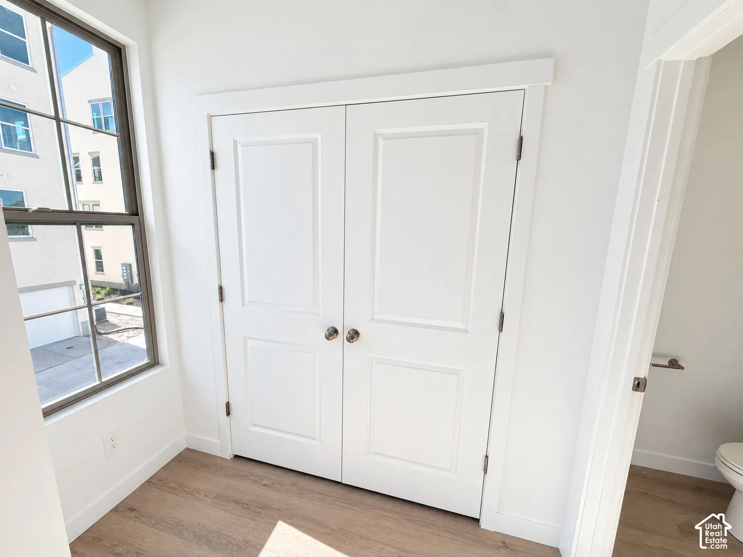 White double-panel closet doors featuring brushed metallic knobs, wood-finish flooring, and a window with a dark-finish frame