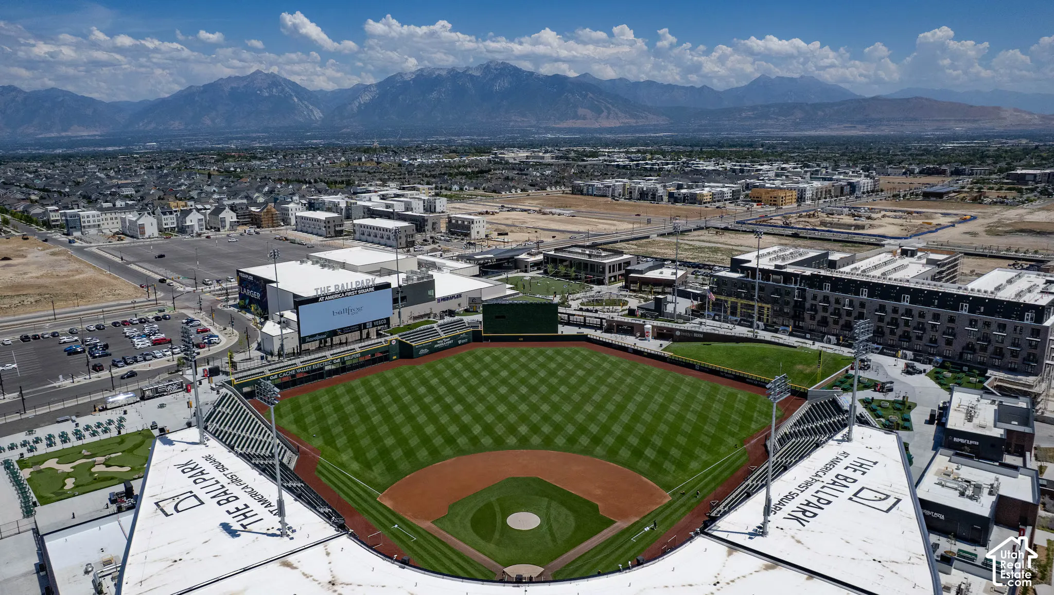Aerial perspective of a baseball stadium featuring a manicured turf field, tiered seating, stadium lighting, and a mountainous backdrop