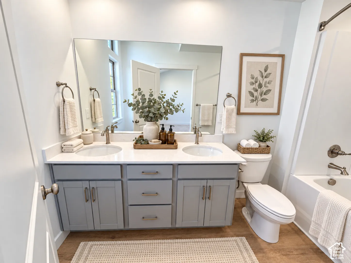 Double vanity featuring light gray cabinetry, solid surface countertop with dual undermount sinks, satin nickel finish hardware, large wall-mounted mirror, and wood-finish flooring