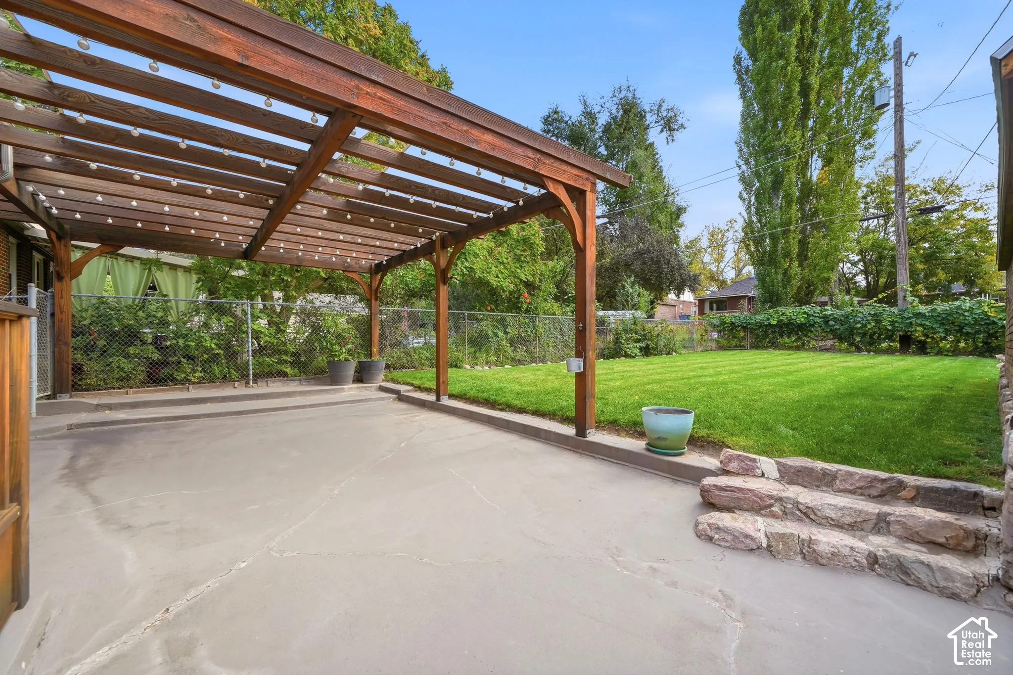 Backyard featuring a wood pergola, concrete patio, natural stone steps, and a grass lawn enclosed by chain-link fencing