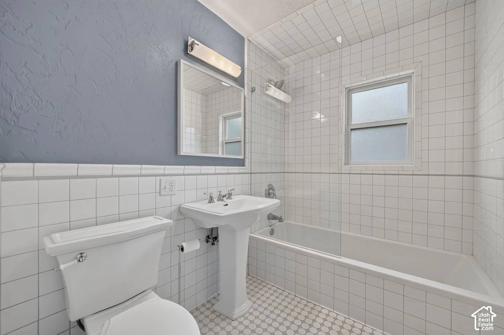 Full bathroom featuring white square wall tile, porcelain pedestal sink, tub-and-shower combination with a glass panel, and light-toned mosaic floor tile