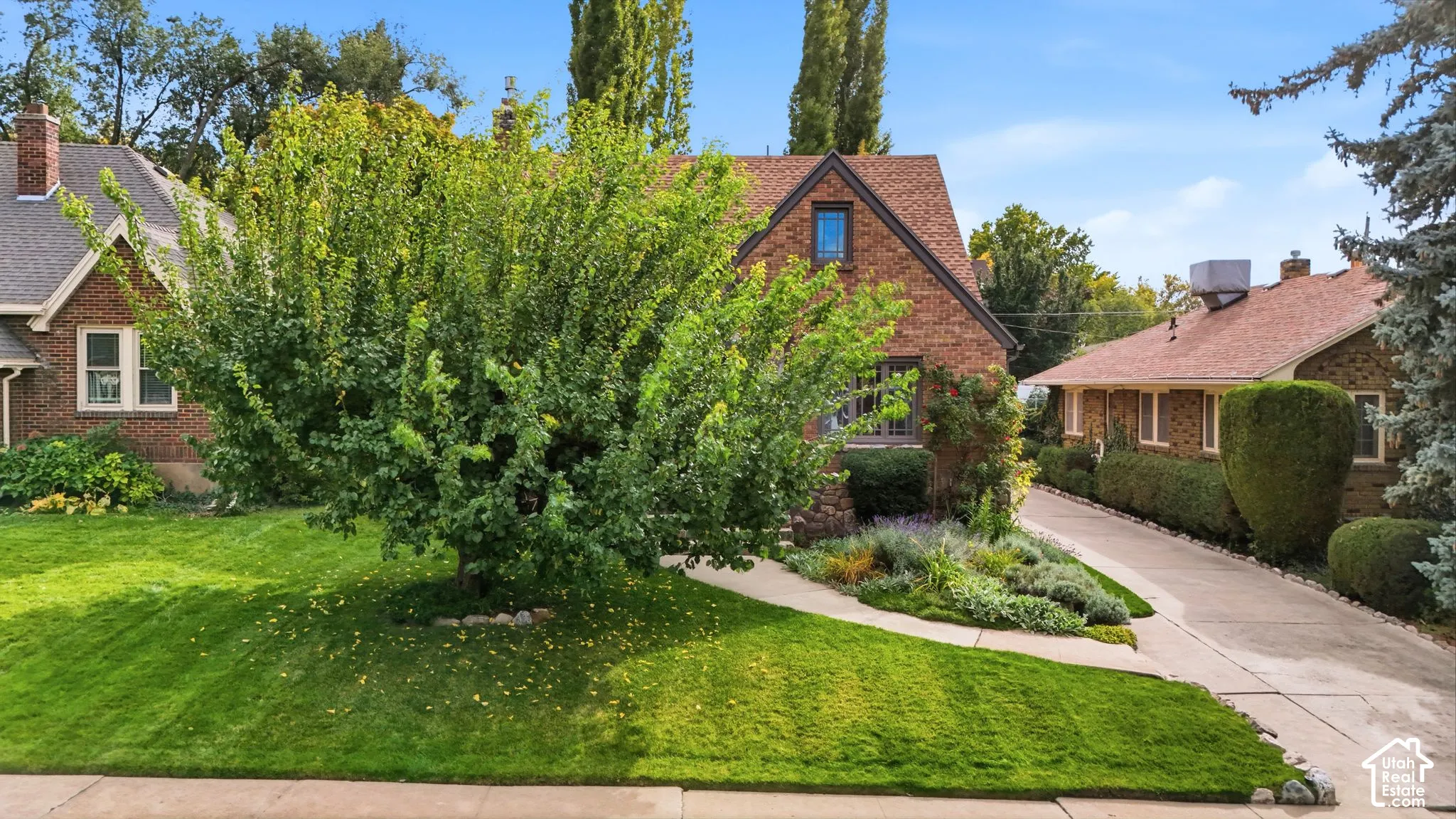 Brick exterior with a gabled roofline, concrete driveway, and landscaped front lawn featuring mature trees and established plantings