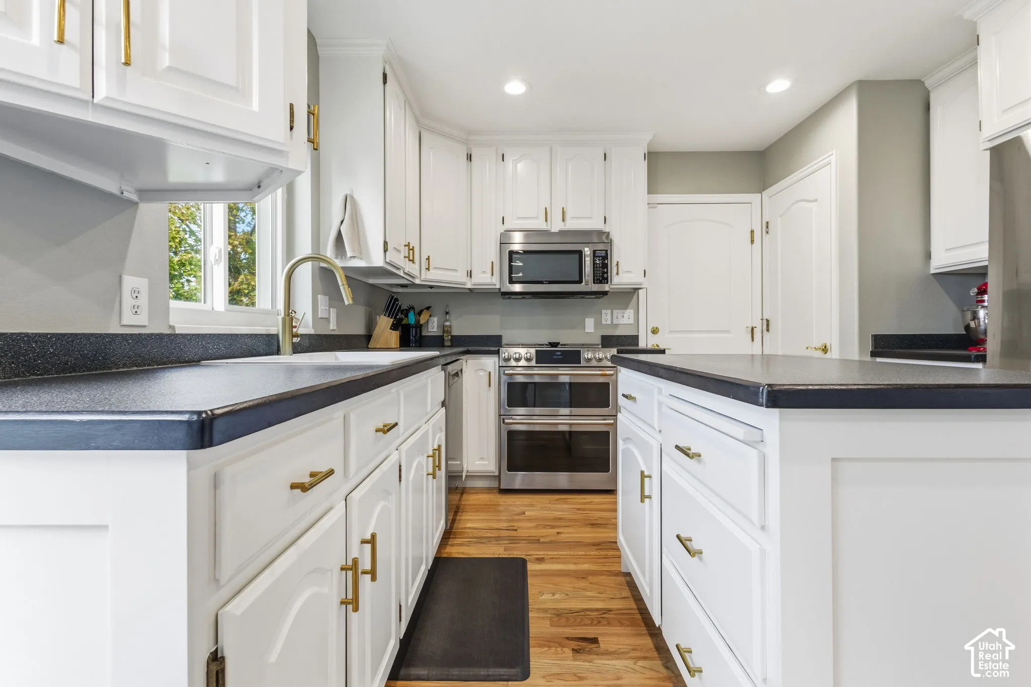 Kitchen featuring white shaker-style cabinetry, gold-tone hardware, stainless steel appliances, a center island with a dark countertop, and wood-finish flooring