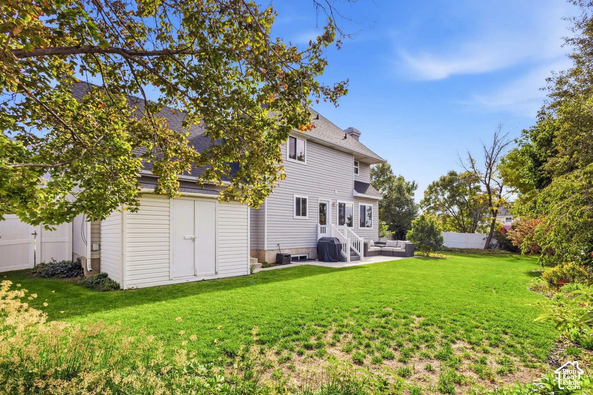 Expansive backyard featuring a level lawn, mature trees, a concrete patio, and white vinyl perimeter fencing