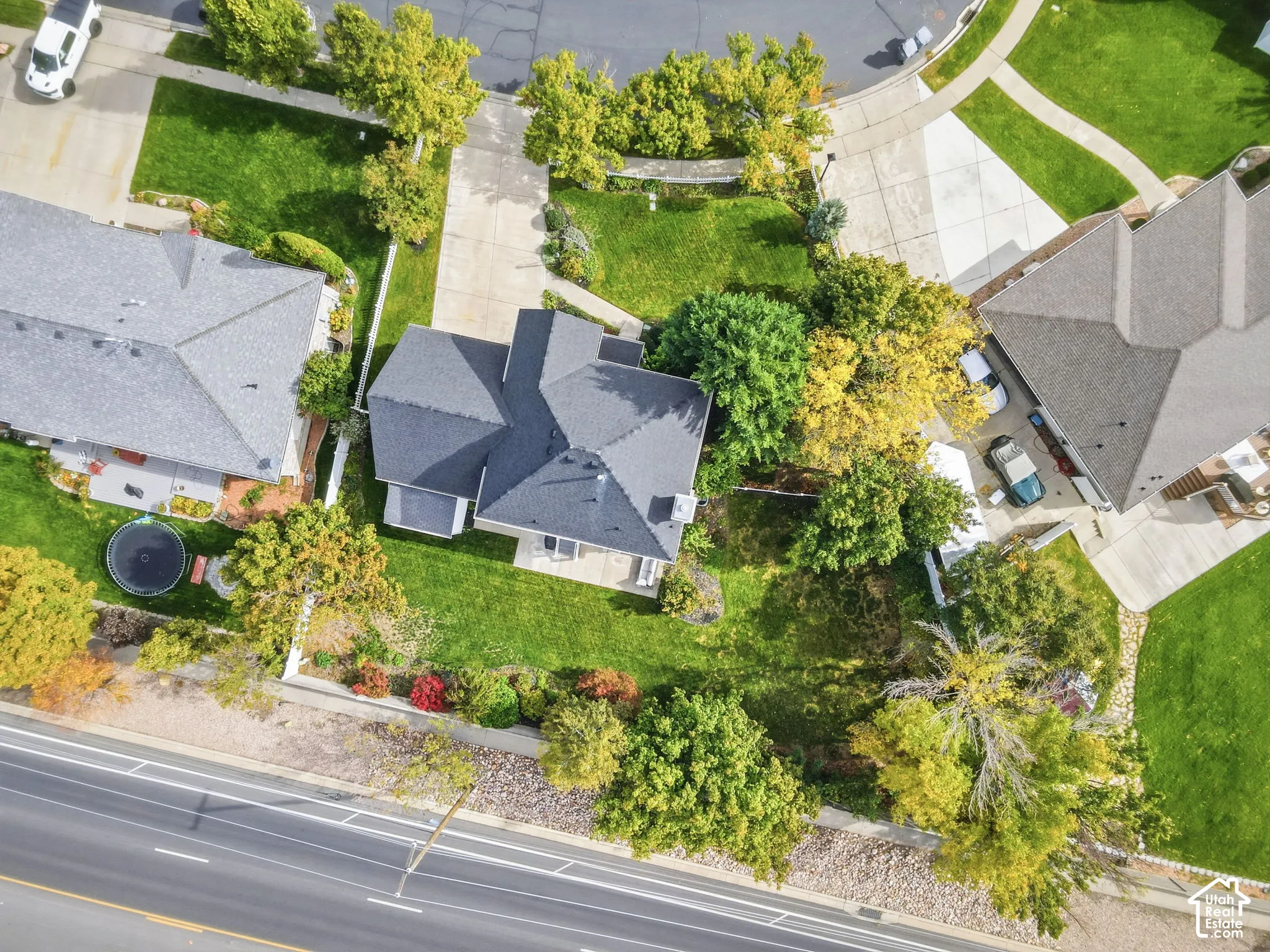 Aerial view of residential property featuring a composition shingle roof, paved driveway, expansive lawn, mature landscaping, and stone hardscaping elements