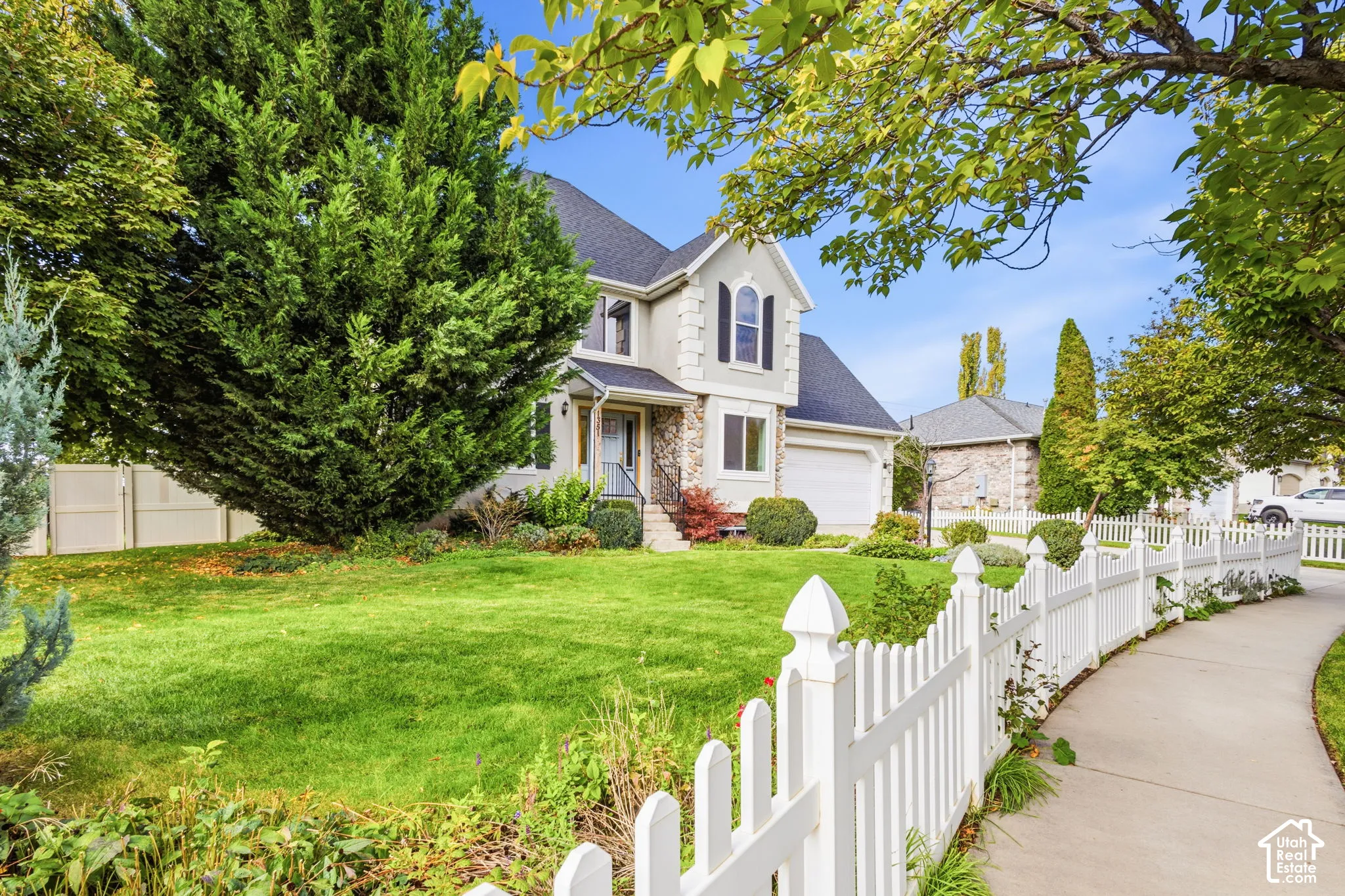 Two-story exterior featuring light-toned siding with stone masonry accents, a shingled roof, an attached two-car garage, and a white picket fence bordering the front lawn