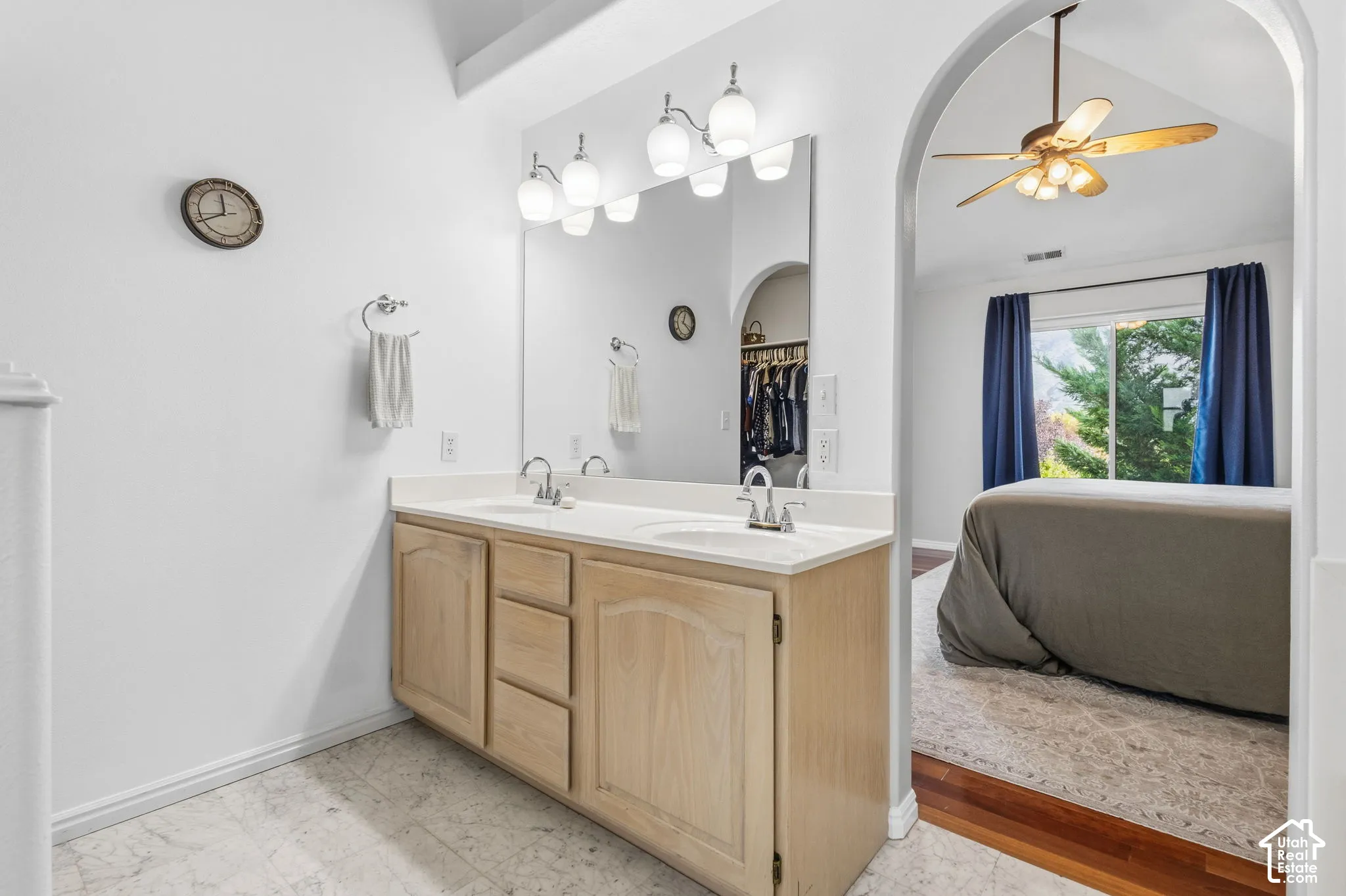 Dual-sink vanity with light-tone wood cabinetry, solid surface countertop, wide-span wall mirror, and multi-bulb vanity lighting