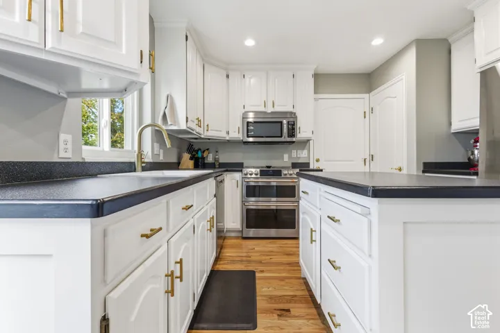 Kitchen featuring white shaker-style cabinetry, gold-tone hardware, stainless steel appliances, a center island with a dark countertop, and wood-finish flooring