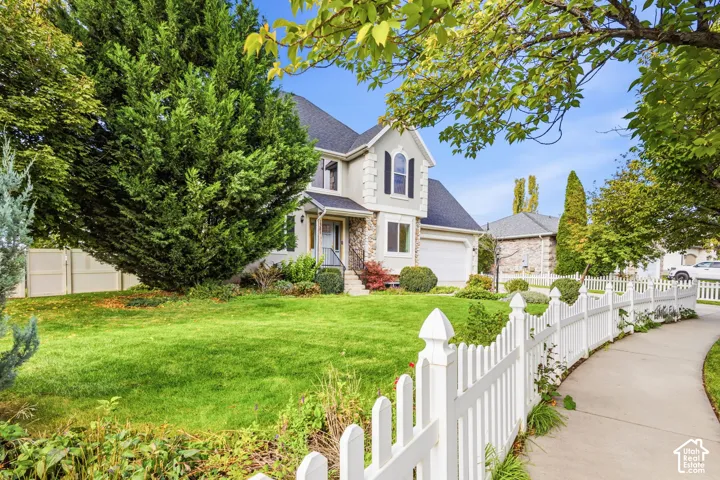 Two-story exterior featuring light-toned siding with stone masonry accents, a shingled roof, an attached two-car garage, and a white picket fence bordering the front lawn