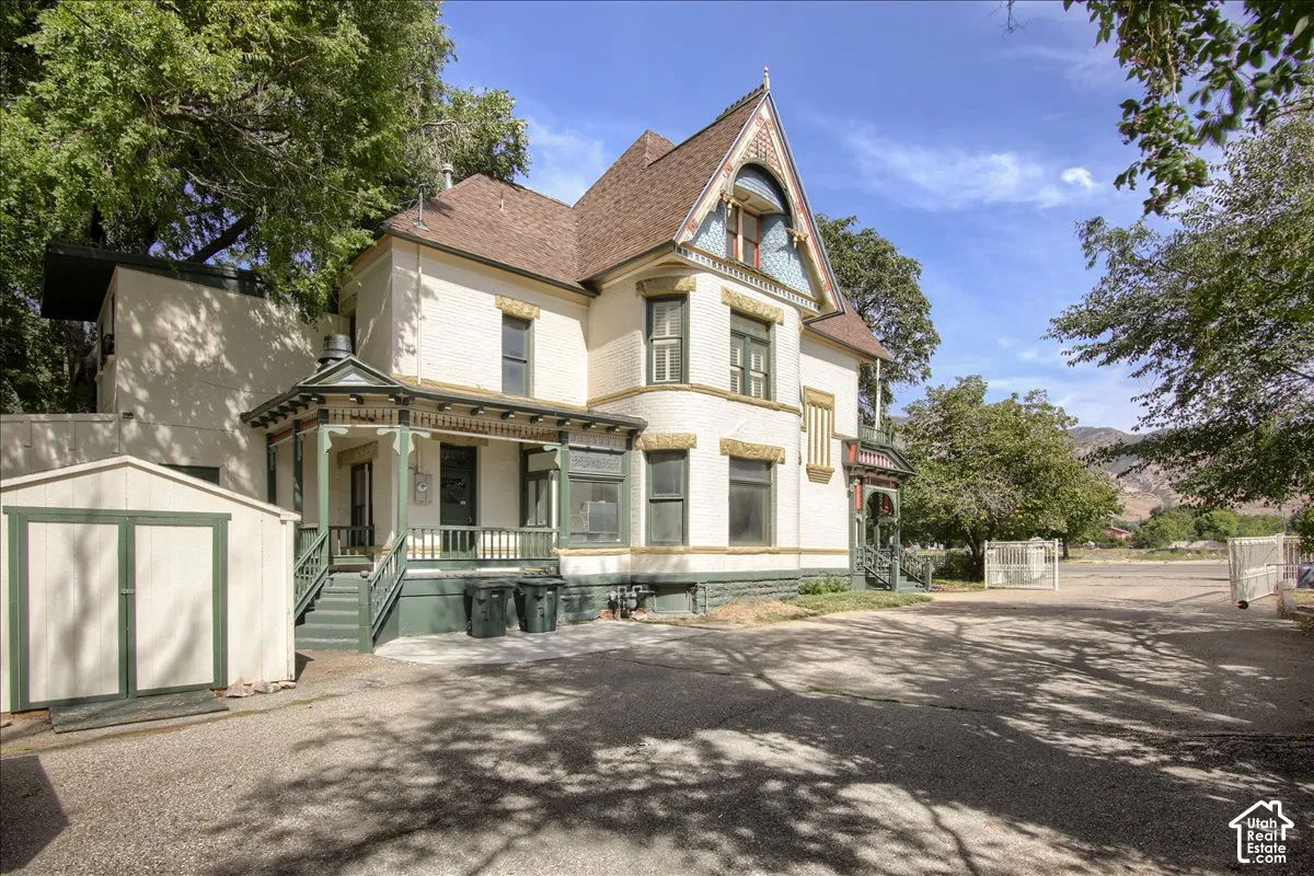 Victorian-style architecture with intricate gable detailing, expansive paved parking area, covered porch with detailed balusters, and a separate storage shed