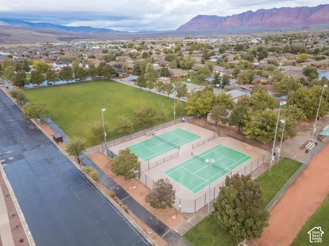 Aerial perspective featuring dual fenced tennis courts, expansive grass area, mature landscaping, and mountain backdrop