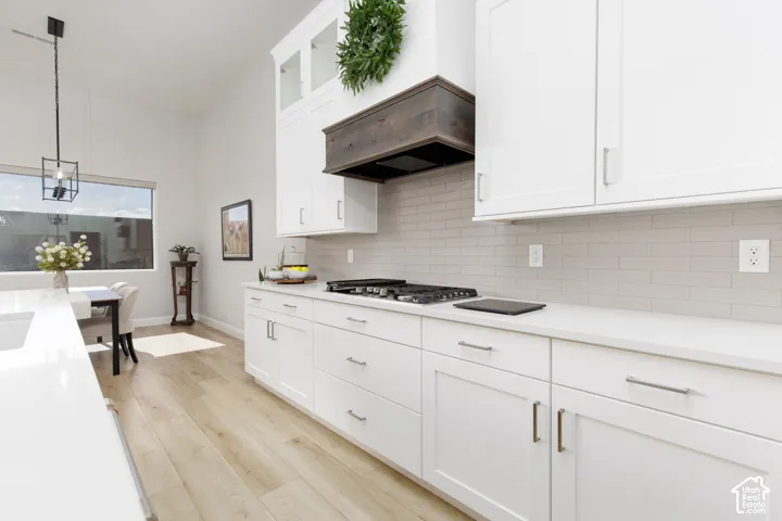 Kitchen featuring white shaker-style cabinetry, a custom dark wood range hood, quartz countertops, a horizontal tile backsplash, and light wood-finish flooring
