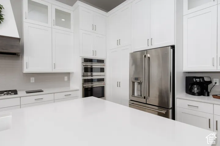 Kitchen featuring ceiling-height shaker-style cabinetry, a solid-surface island, stainless steel double wall ovens, a stainless steel refrigerator, and a tiled backsplash