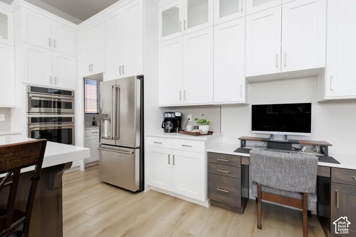 Kitchen featuring floor-to-ceiling white cabinetry with glass-front uppers, stainless steel double wall ovens, built-in desk workstation, quartz countertops, and wood-finish flooring